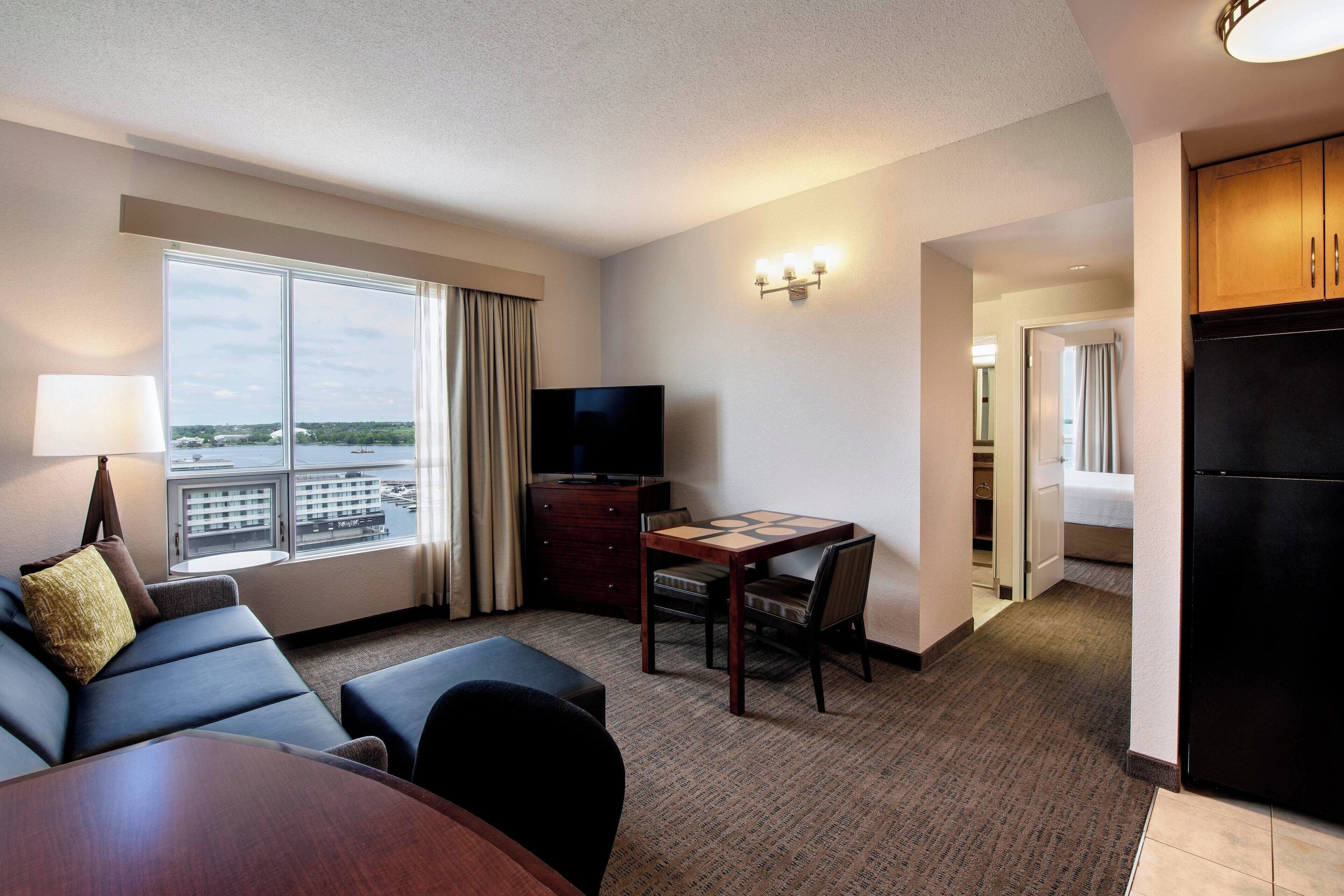 A hotel room with a large window, a flat-screen TV, a dining area, and a kitchenette.