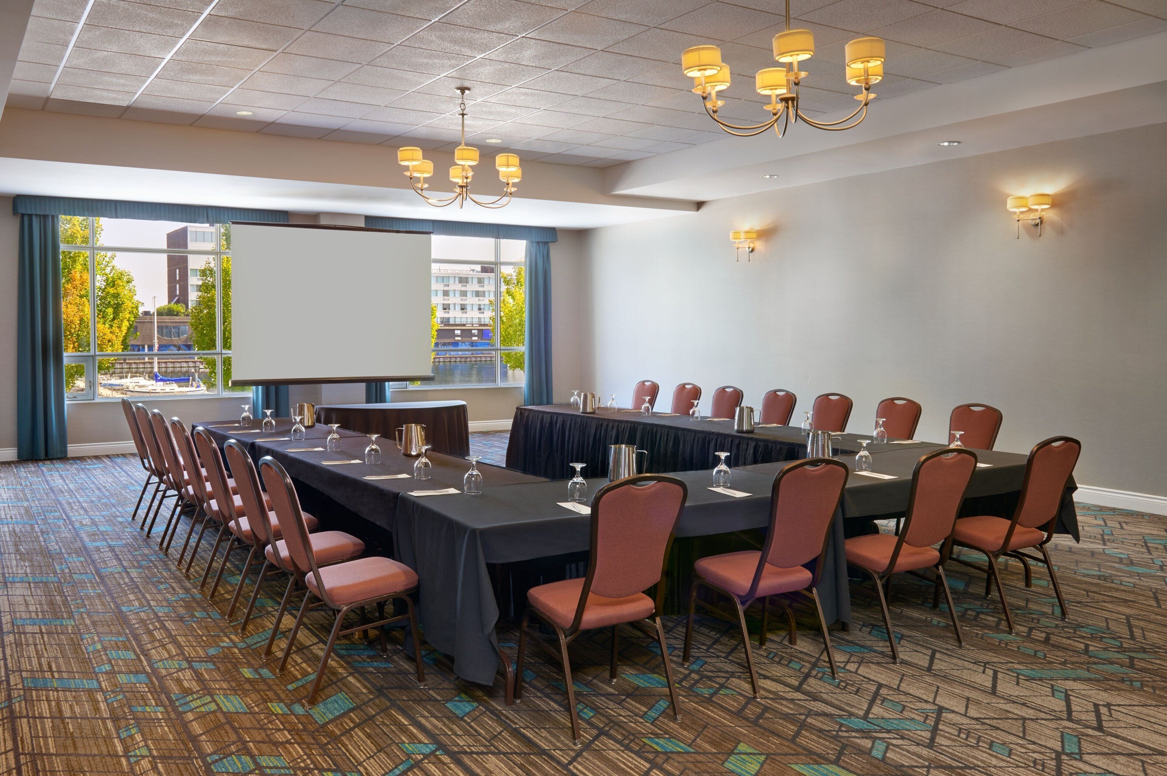 A conference room with a long table, chairs, a projector screen, and a view of buildings through large windows.