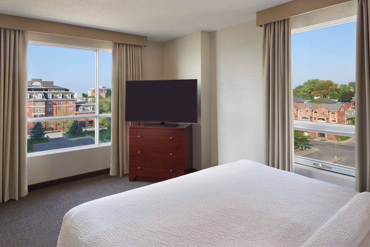 A hotel room with a large bed, a dresser, a flat-screen TV, and a view of buildings through the windows.