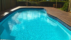 Una piscina rectangular con borde de azulejos, rodeada por un suelo de ladrillo y una cerca de metal.