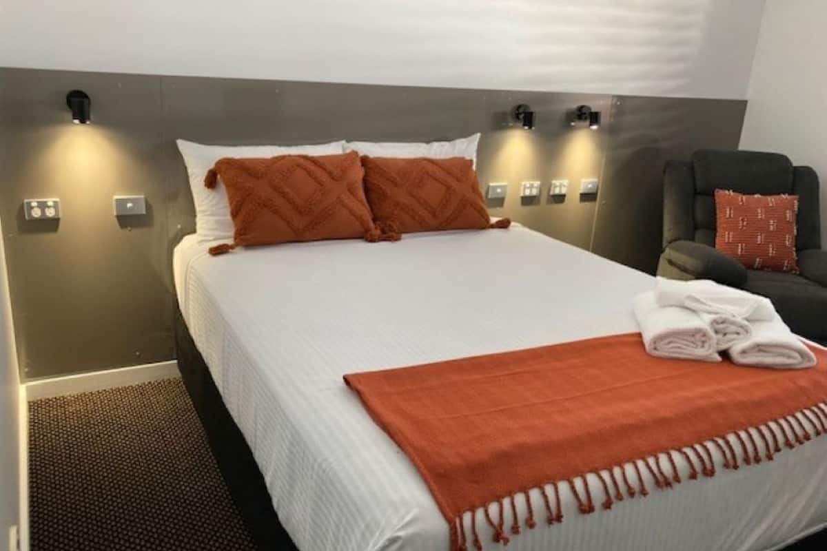 A neatly made bed with a white and orange blanket, two pillows, and a folded towel on the side.