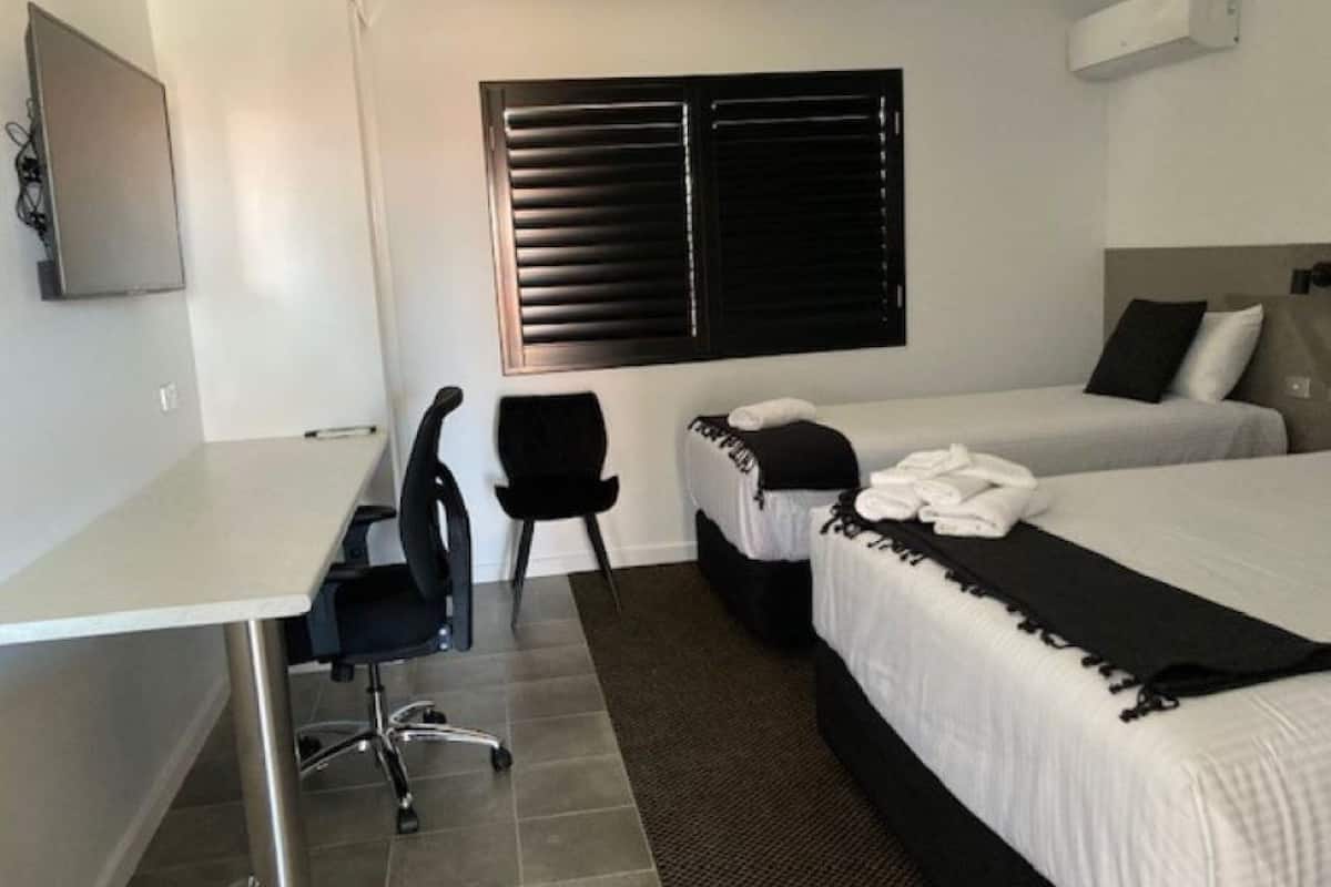 A hotel room with a desk, two beds, a chair, and a window with blinds.