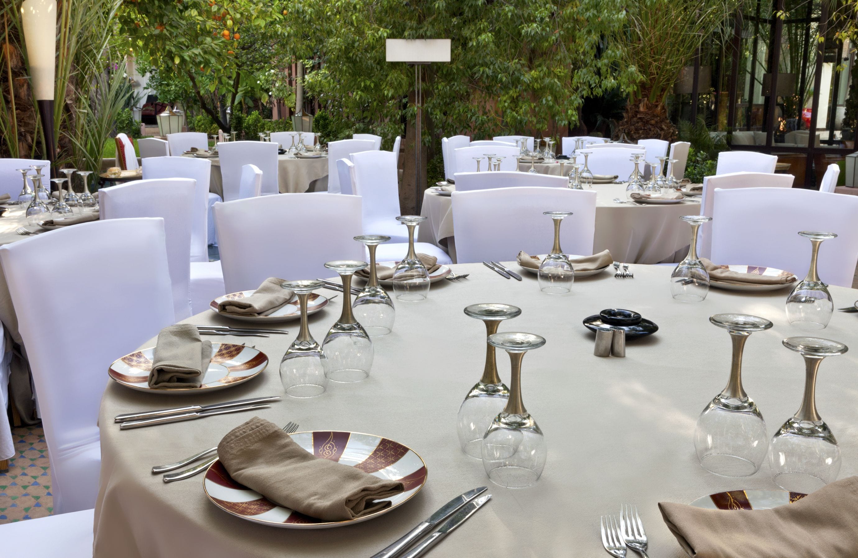 Un espacio comedor exterior formal con mesas redondas dispuestas con sillas blancas, cubiertos y copas de vino.