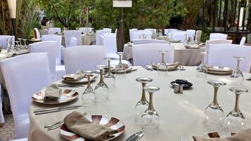 Un espacio comedor exterior formal con mesas redondas dispuestas con sillas blancas, cubiertos y copas de vino.