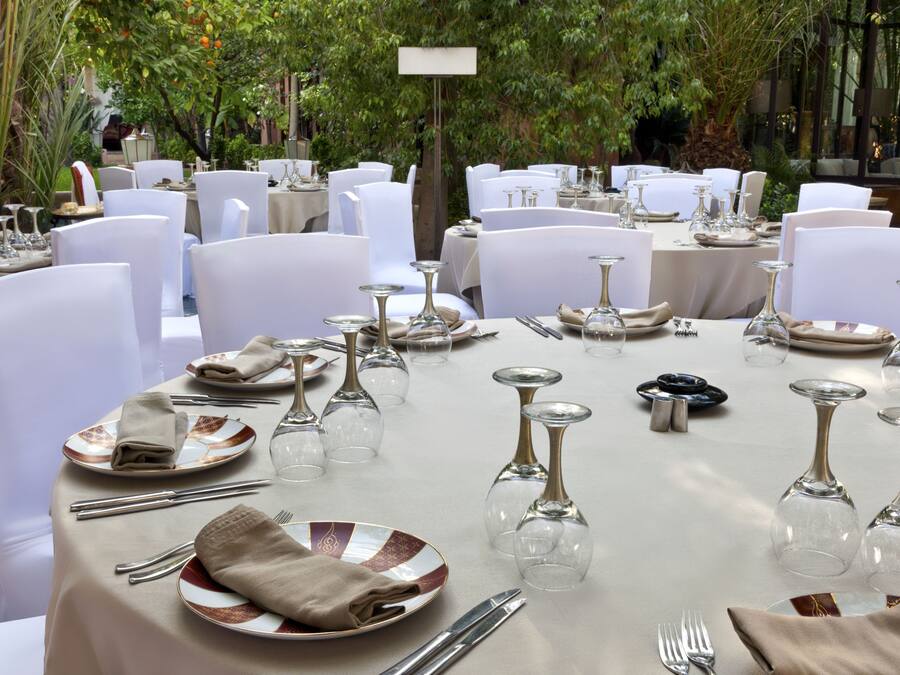Un espacio comedor exterior formal con mesas redondas dispuestas con sillas blancas, cubiertos y copas de vino.