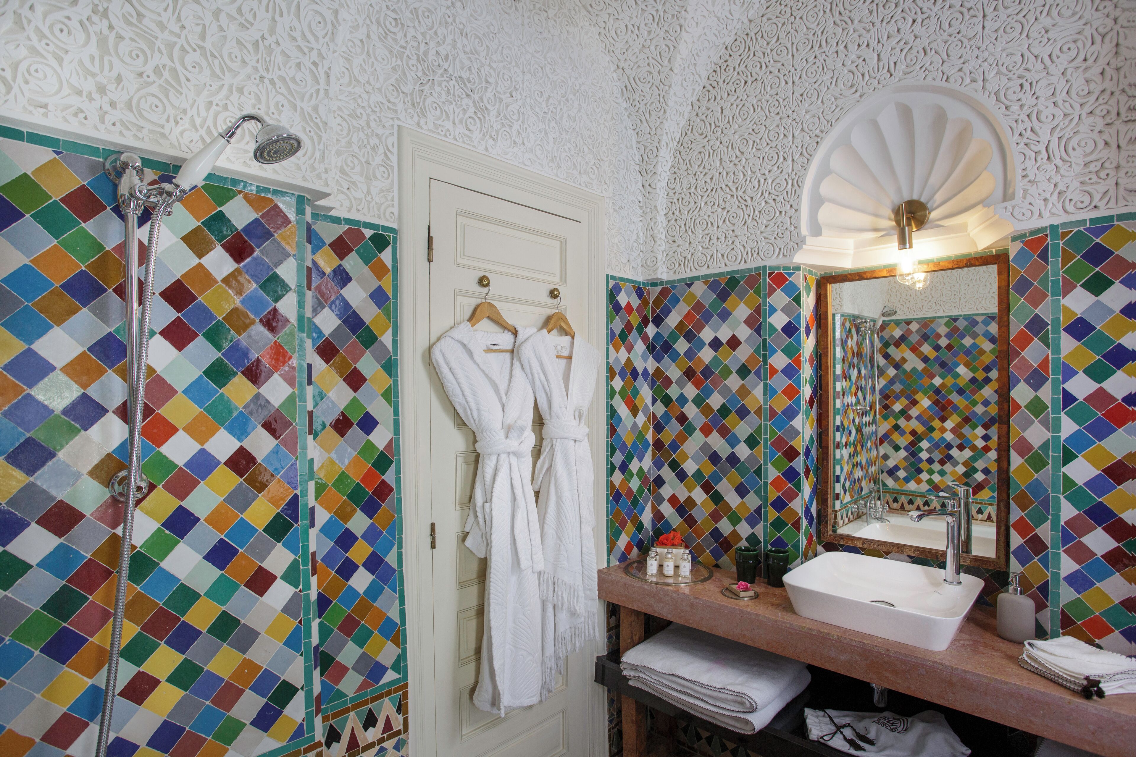 Une salle de bain avec un mur recouvert de carreaux aux motifs géométriques colorés, un lavabo blanc et un miroir surmonté d’un décor en forme d’éventail.