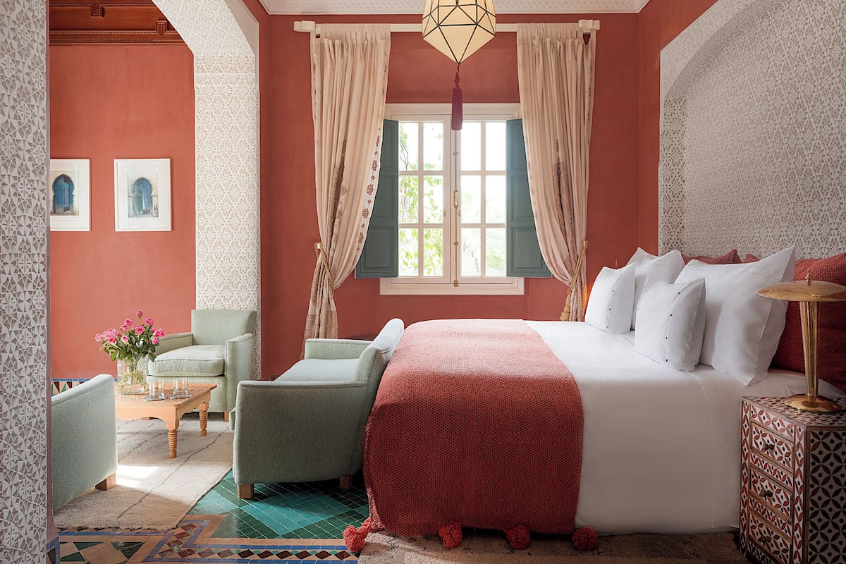 A bedroom with a large bed, two armchairs, a small table with a vase of flowers, and a window with curtains.