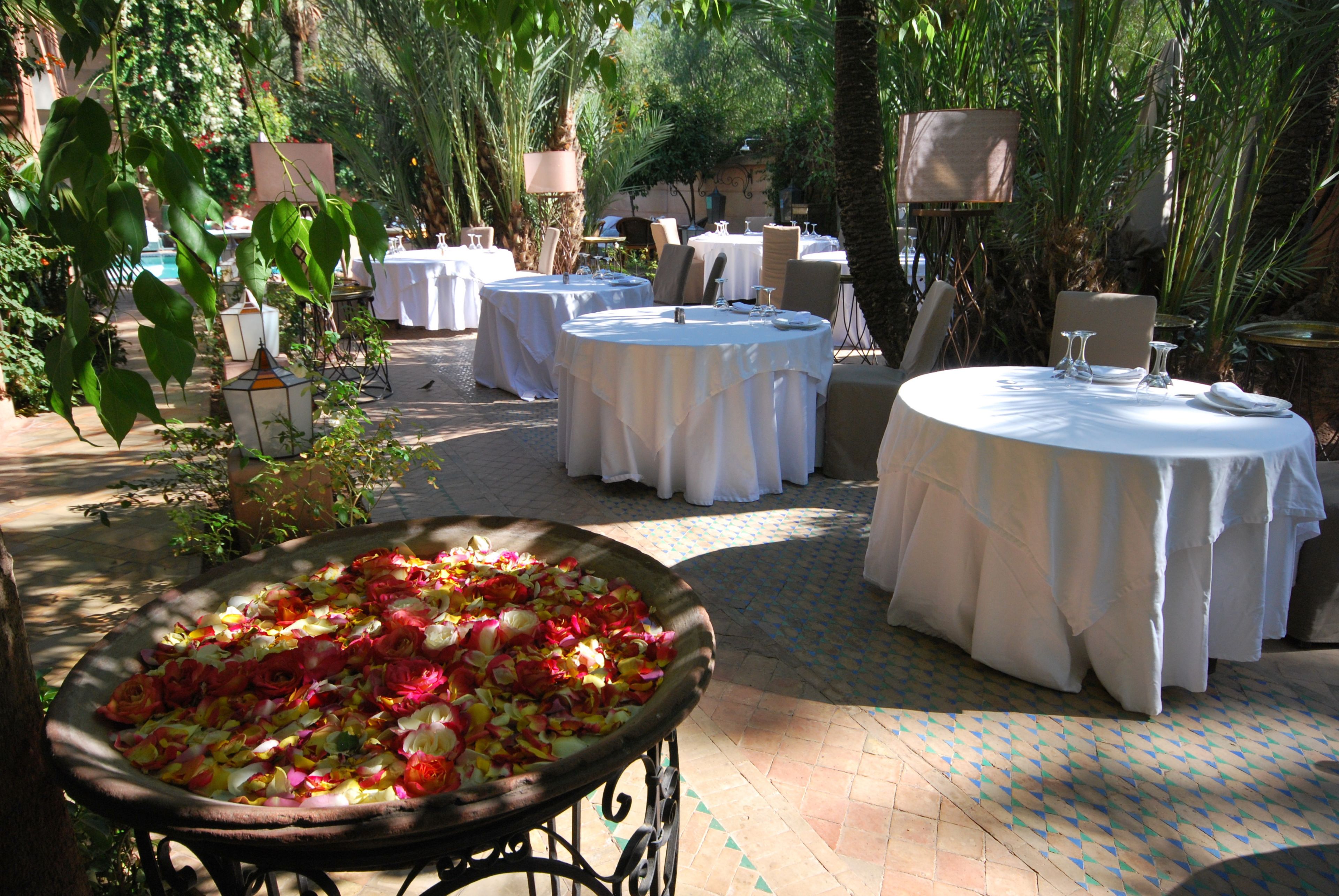 An outdoor dining area with round tables covered in white tablecloths, surrounded by greenery and plants.