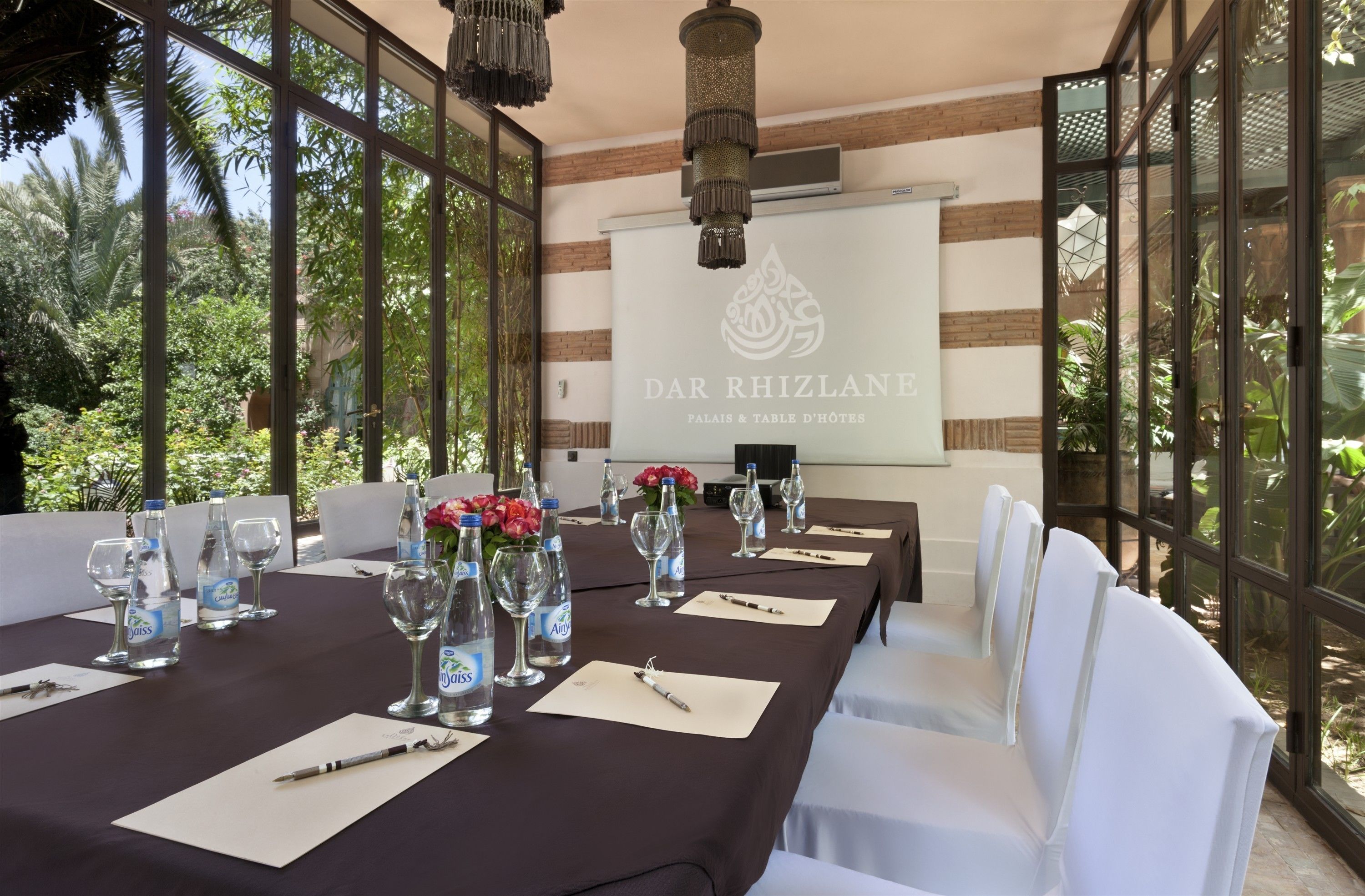 A conference room with a long table set for a meeting, surrounded by white chairs, with a large window offering a view of greenery.