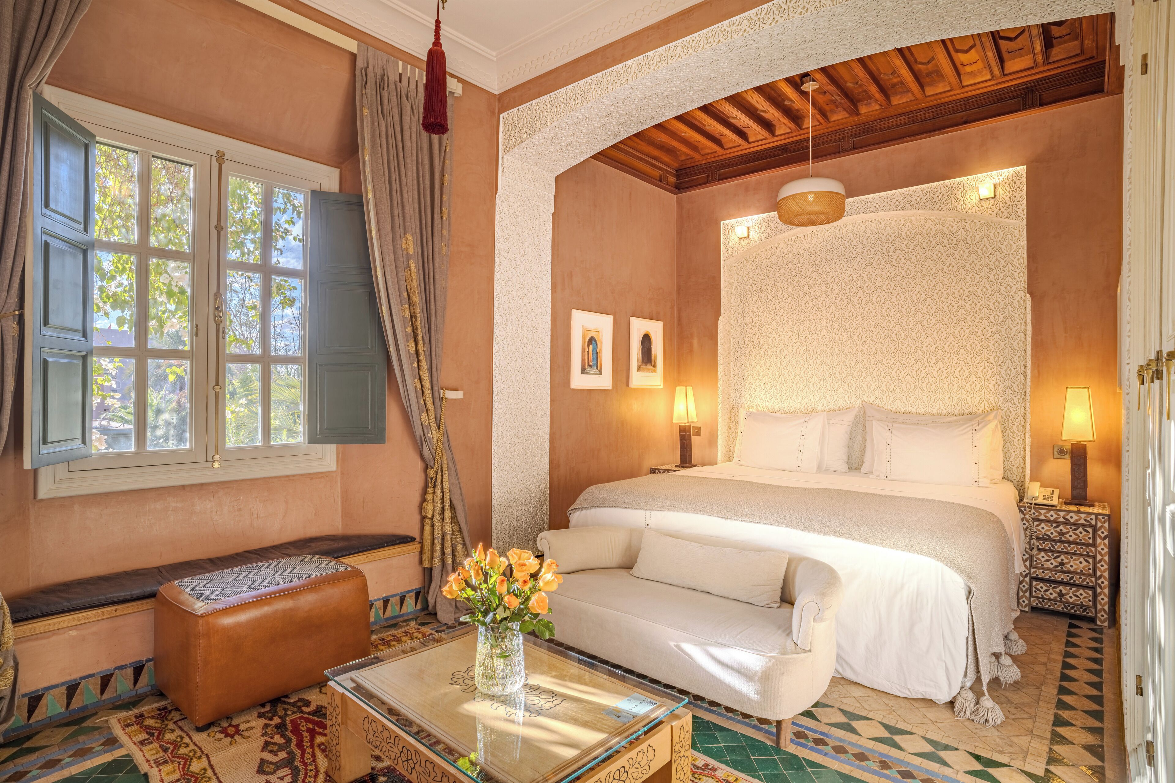 A bedroom with a large bed, a bench, a glass table with a vase, and patterned floor tiles.