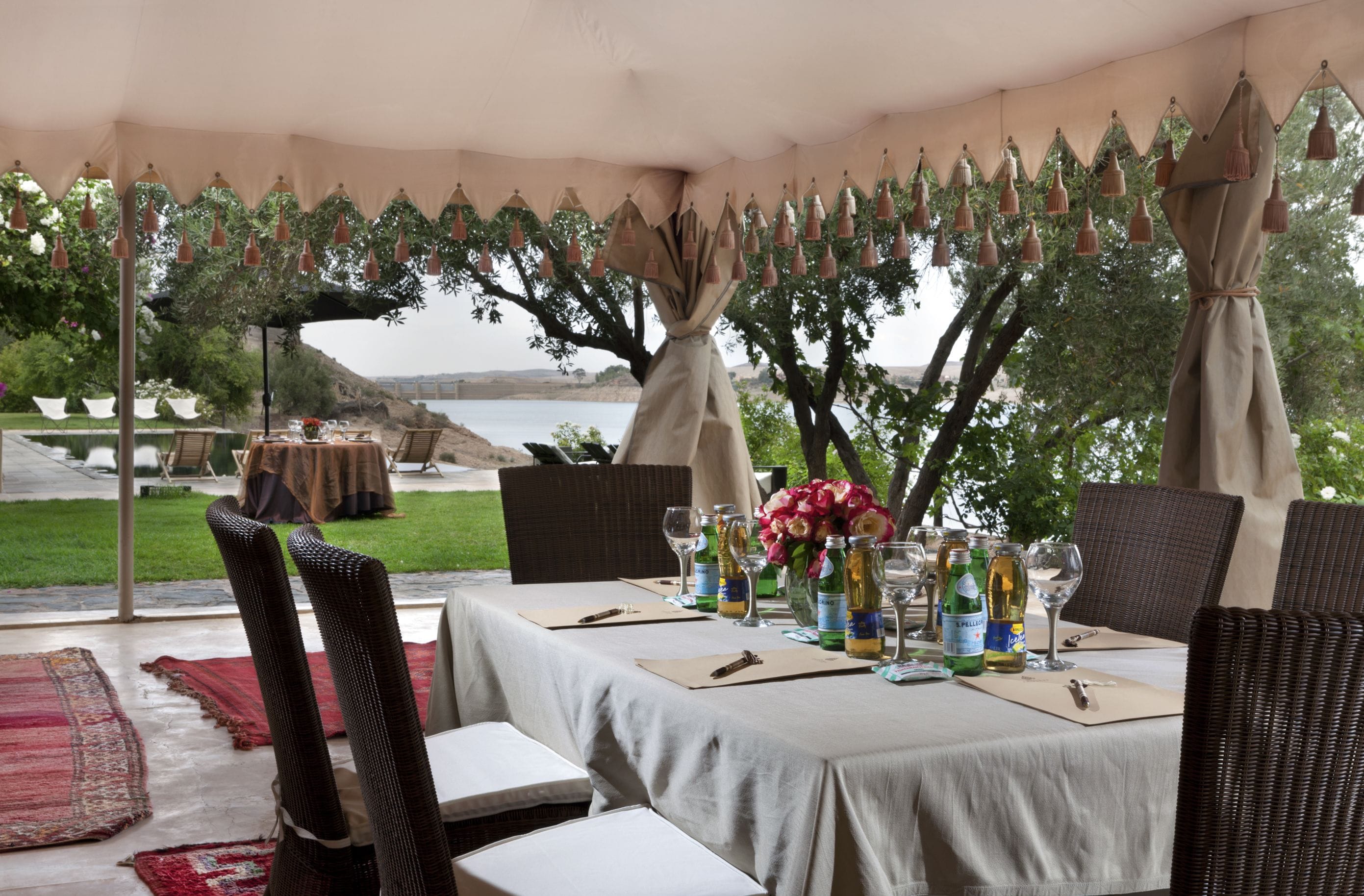 Un área de comedor al aire libre con mesas preparadas para una comida, rodeada de árboles y vistas al agua.