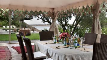 Un área de comedor al aire libre con mesas preparadas para una comida, rodeada de árboles y vistas al agua.
