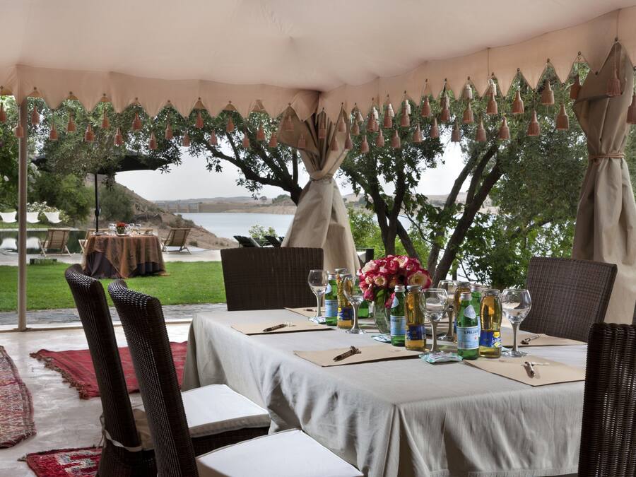 Un área de comedor al aire libre con mesas preparadas para una comida, rodeada de árboles y vistas al agua.