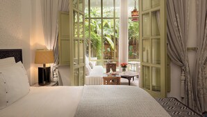 A hotel room with a large bed, a nightstand with a lamp, a small table with a vase of flowers, and a view of greenery outside. - Dar Rhizlane (Marrakech)
