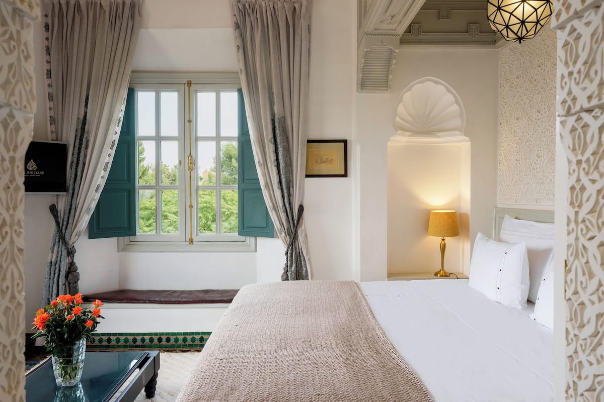 A hotel room with a bed, a nightstand with a lamp, a vase with flowers, and a window with curtains.