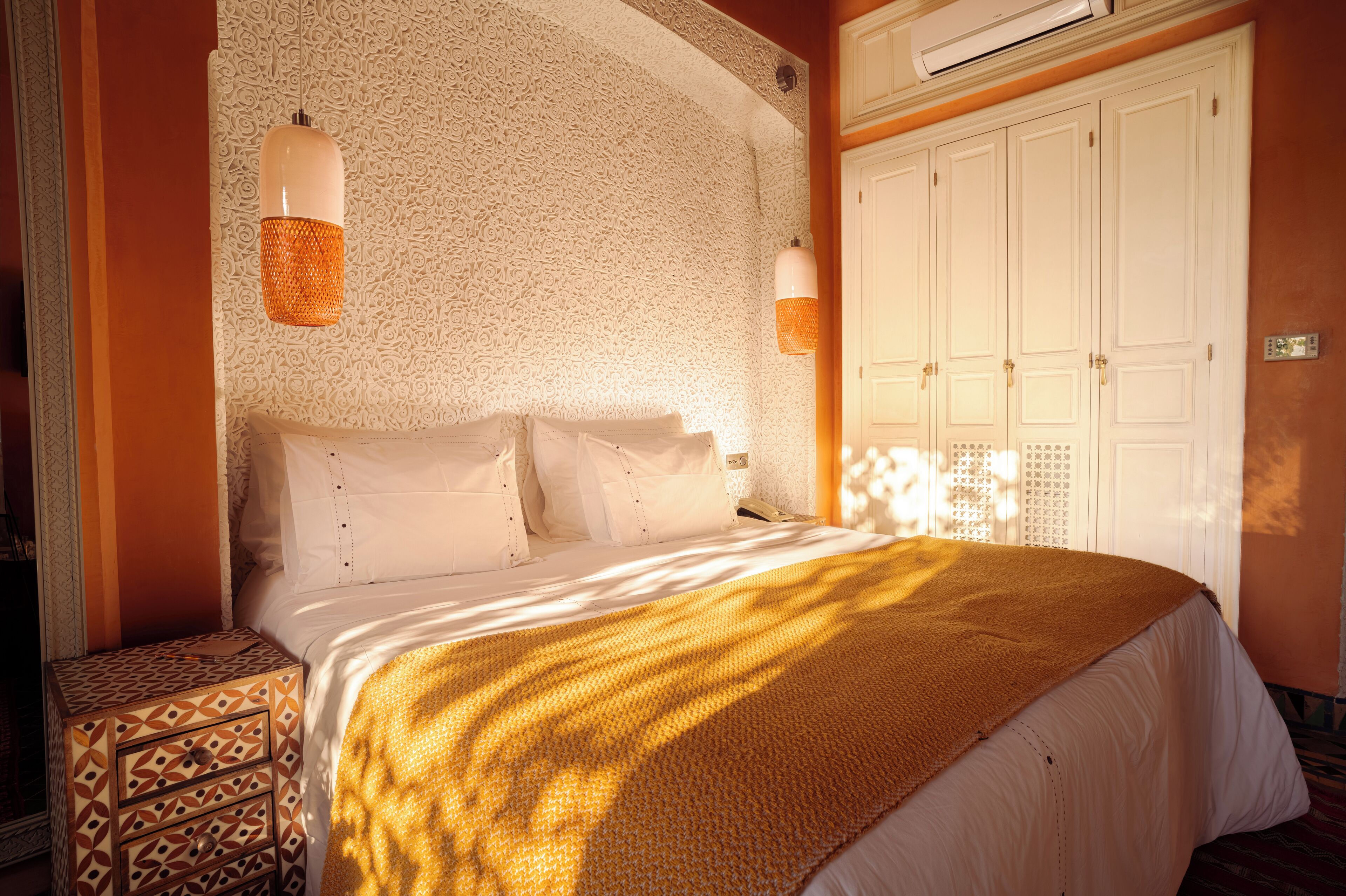 A neatly made bed with a patterned headboard, a bedside table with drawers, and a large white wardrobe.