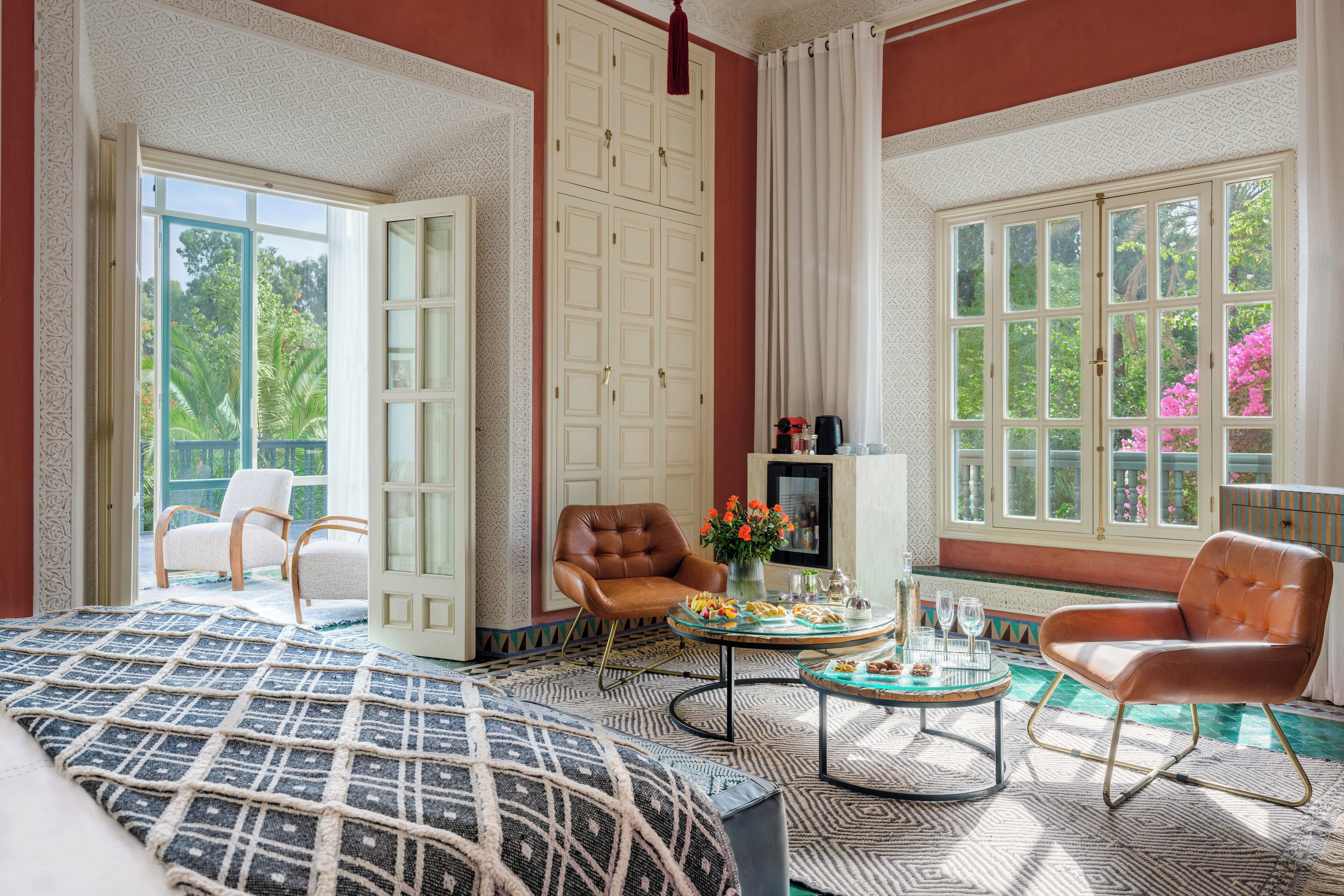 A spacious room with a large bed, two armchairs, a small table with a vase and snacks, and a view of greenery through large windows.