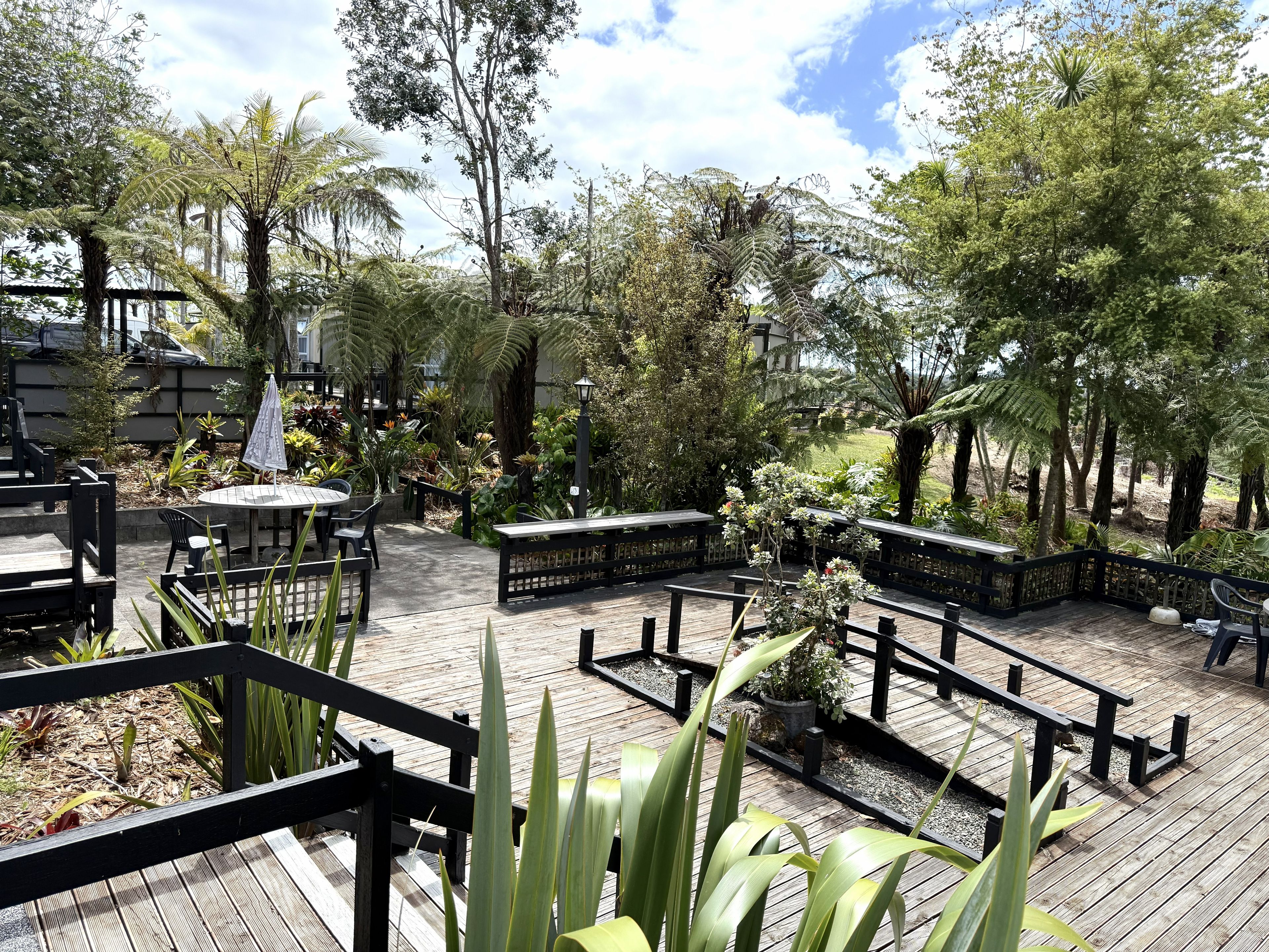 An outdoor patio area with wooden decking, tables, and chairs, surrounded by various plants and trees.