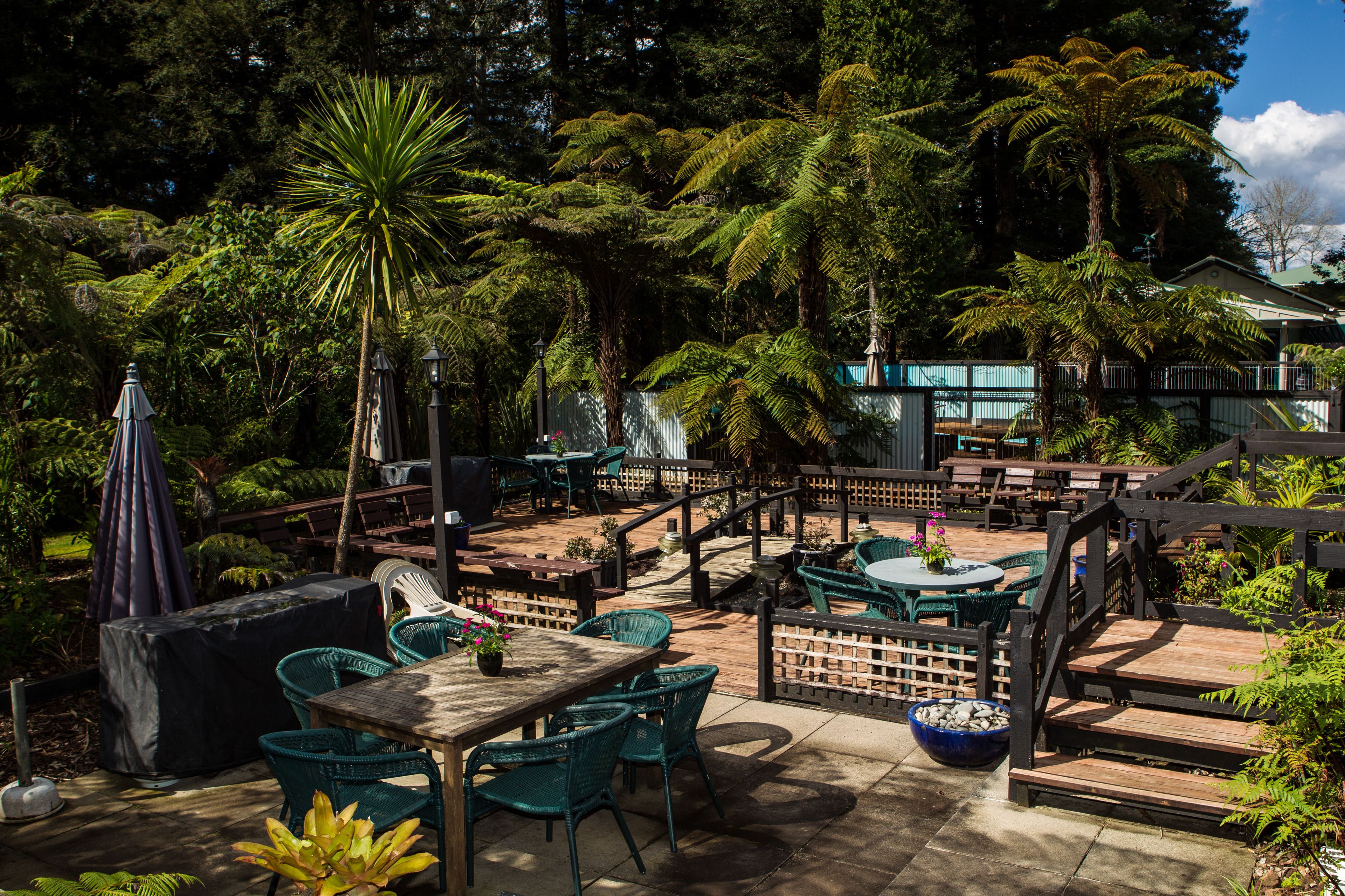An outdoor patio area with wooden tables and chairs, surrounded by lush greenery and a wooden fence.