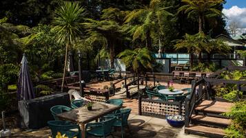 An outdoor patio area with wooden tables and chairs, surrounded by lush greenery and a wooden fence.