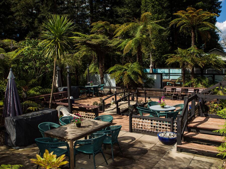 An outdoor patio area with wooden tables and chairs, surrounded by lush greenery and a wooden fence.