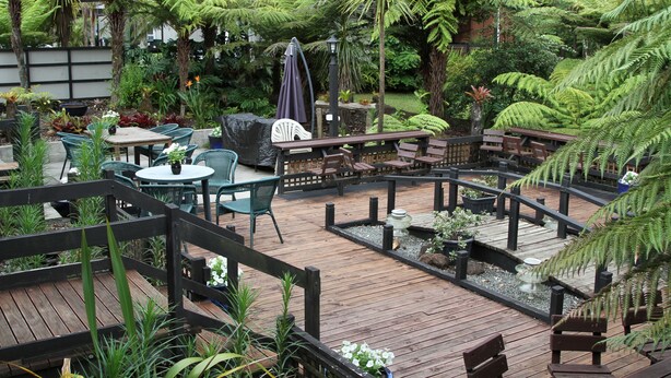 Um deck de madeira com mobiliário externo, cercado por vegetação exuberante e uma pequena cascata.