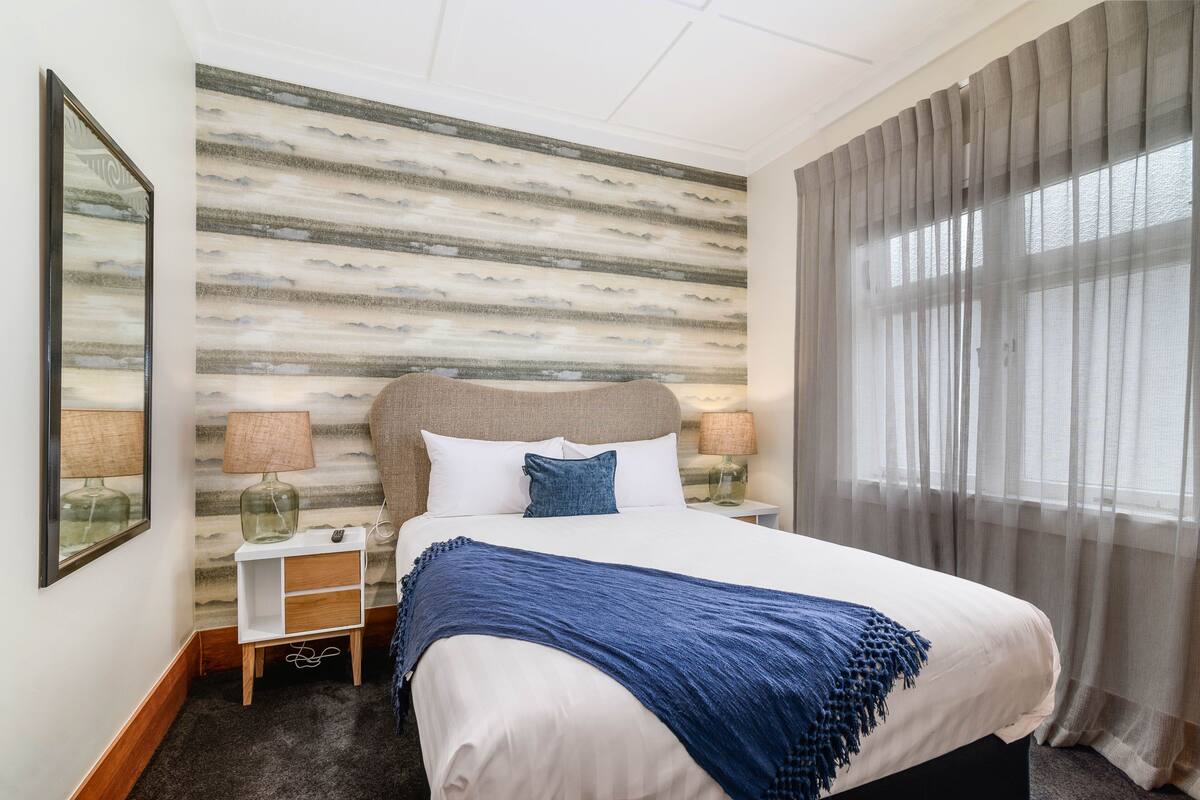 A neatly arranged bedroom with a bed, bedside tables, lamps, and a patterned wall.