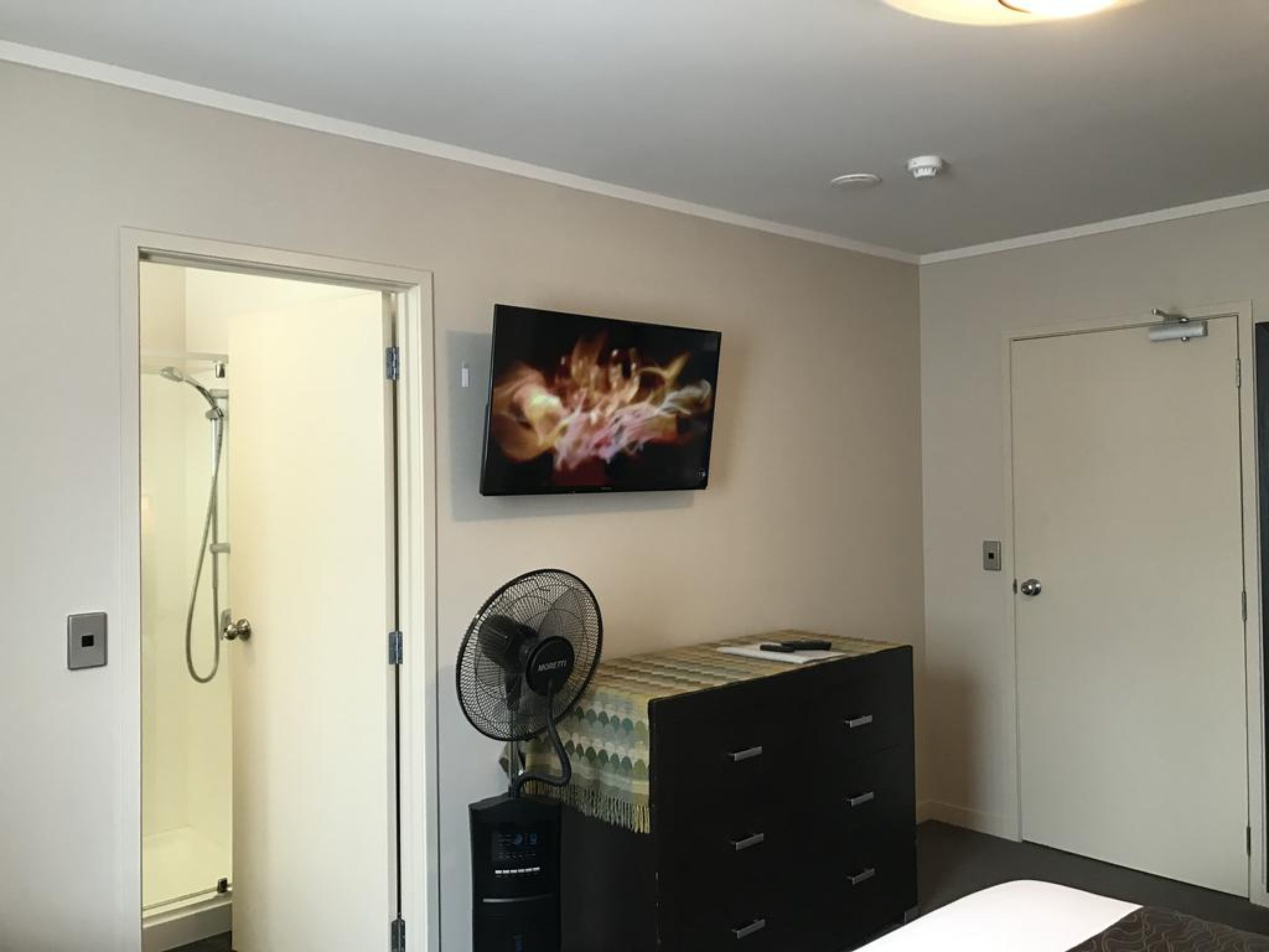 A bathroom with a shower, a mounted TV, a fan, and a dresser.