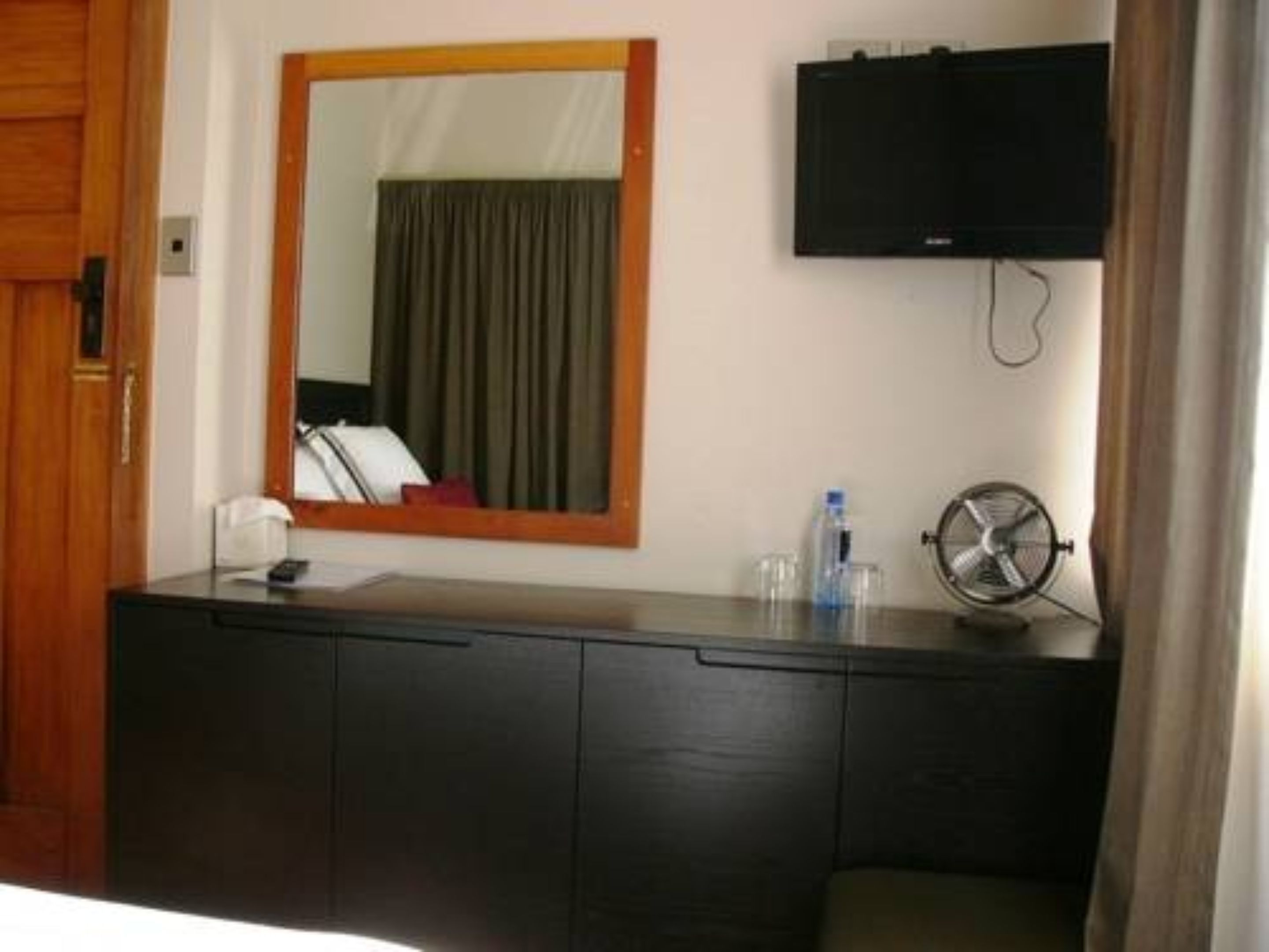 Habitación de hotel con una puerta de madera, un aparador de madera oscura, un espejo, un televisor de pantalla plana, un pequeño ventilador y una botella de agua sobre el aparador.