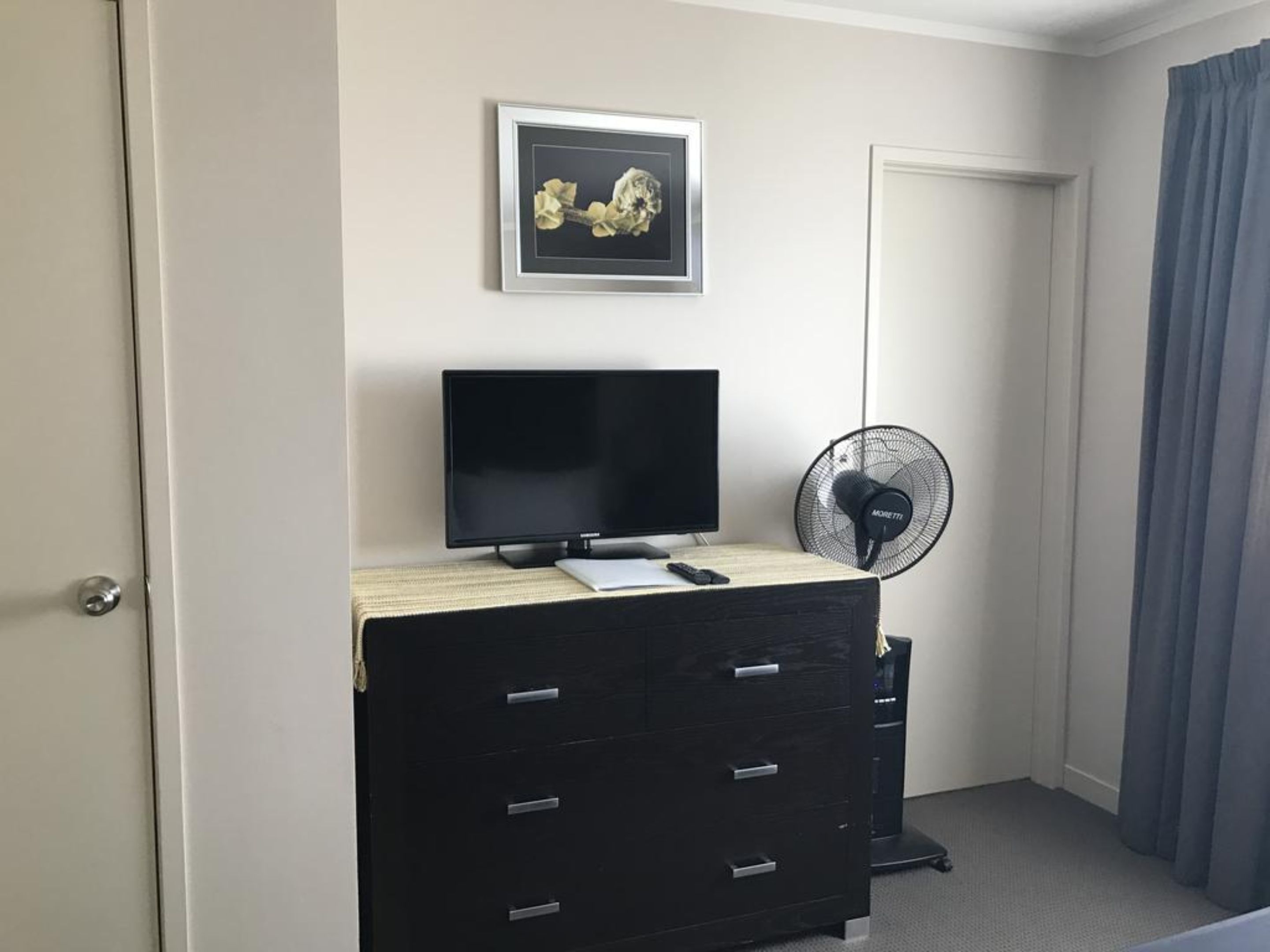 Une pièce avec une commode noire, un téléviseur à écran plat, un tableau encadré au mur, un ventilateur sur pied et une porte avec un rideau.
