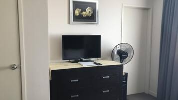 Une pièce avec une commode noire, un téléviseur à écran plat, un tableau encadré au mur, un ventilateur sur pied et une porte avec un rideau.