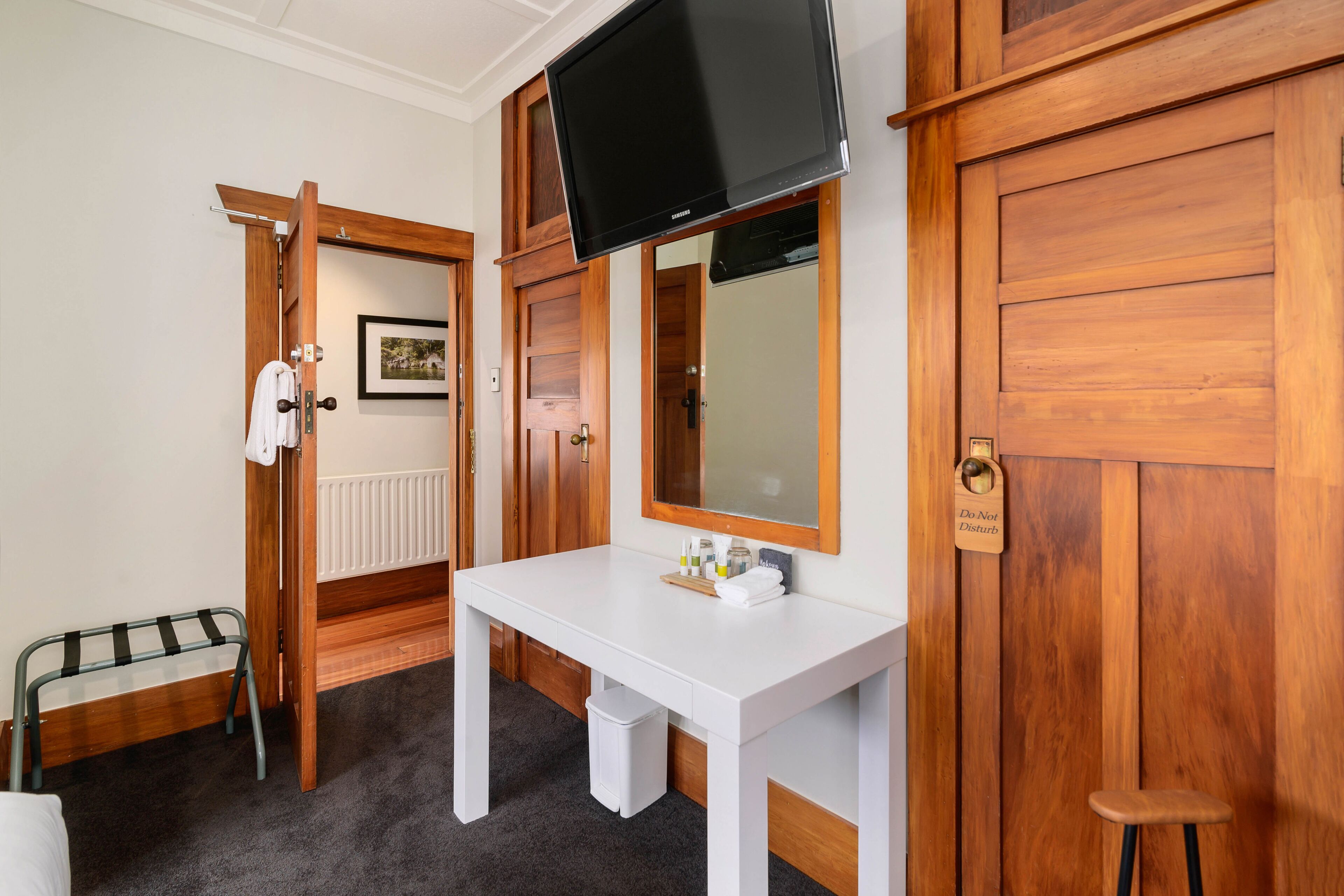 A hotel room with a white desk, a television mounted above it, a wooden wardrobe, a door with a do not disturb sign, and a small stool.