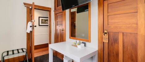 Habitación de hotel con un escritorio blanco, un televisor montado encima, un armario de madera, una puerta con un cartel de ‘no molestar’ y un taburete pequeño.