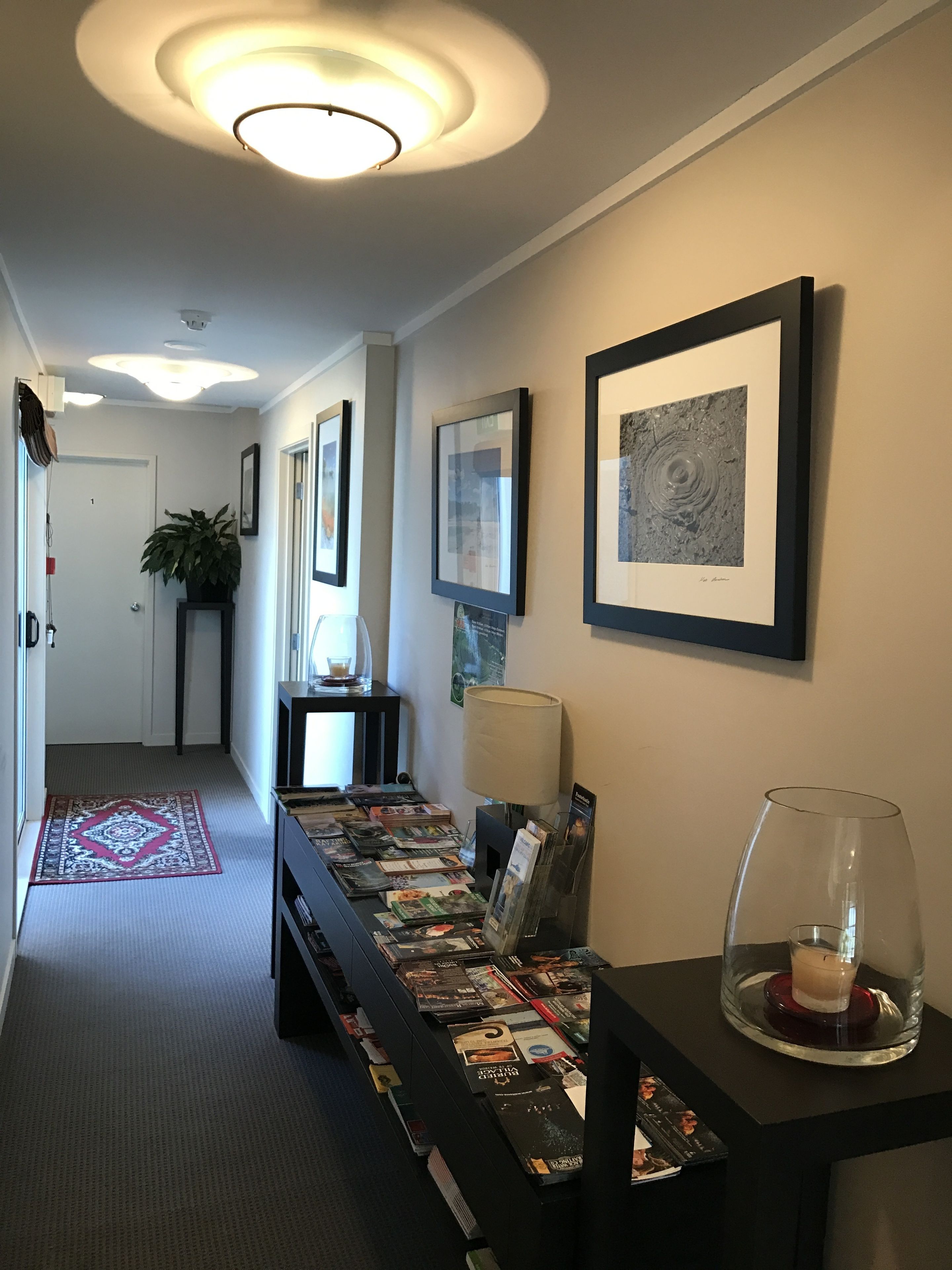 A hallway with framed artwork on the walls, a desk with magazines, a lamp, and a glass dome with a candle.