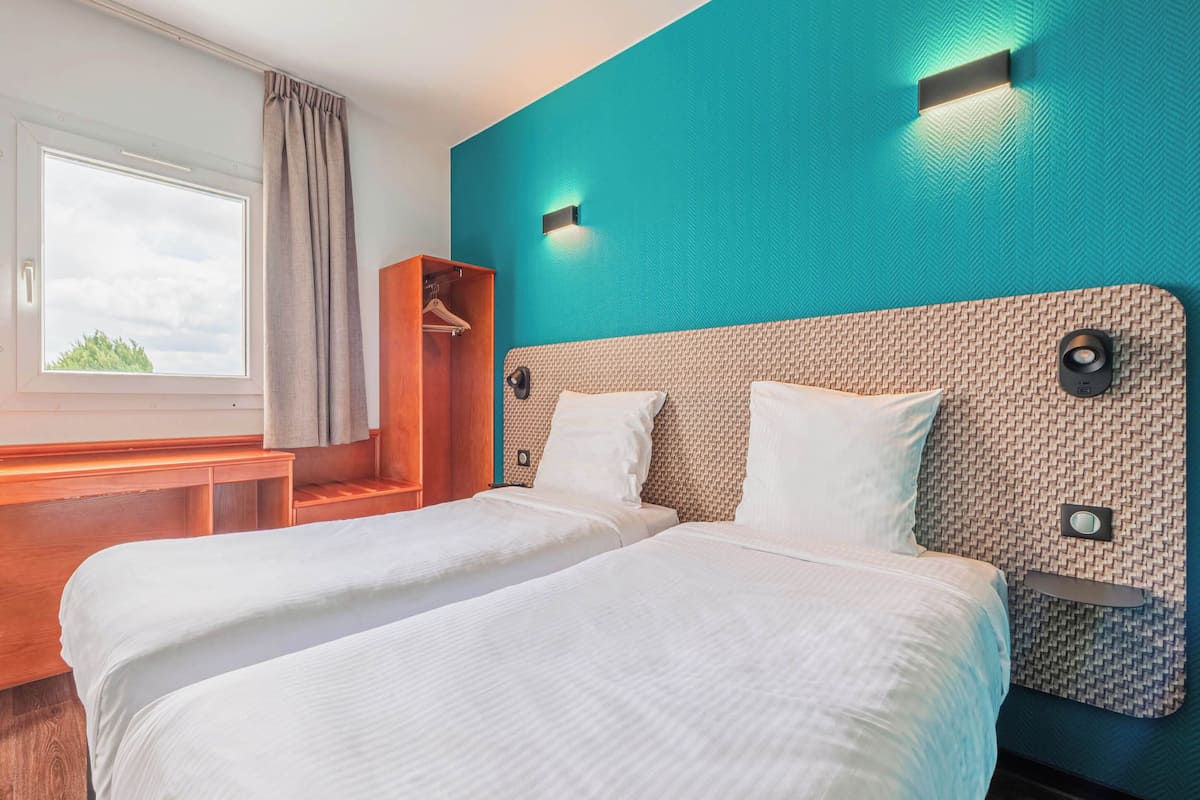 Une chambre d’hôtel avec deux lits, un banc en bois, une armoire et une fenêtre avec des rideaux.