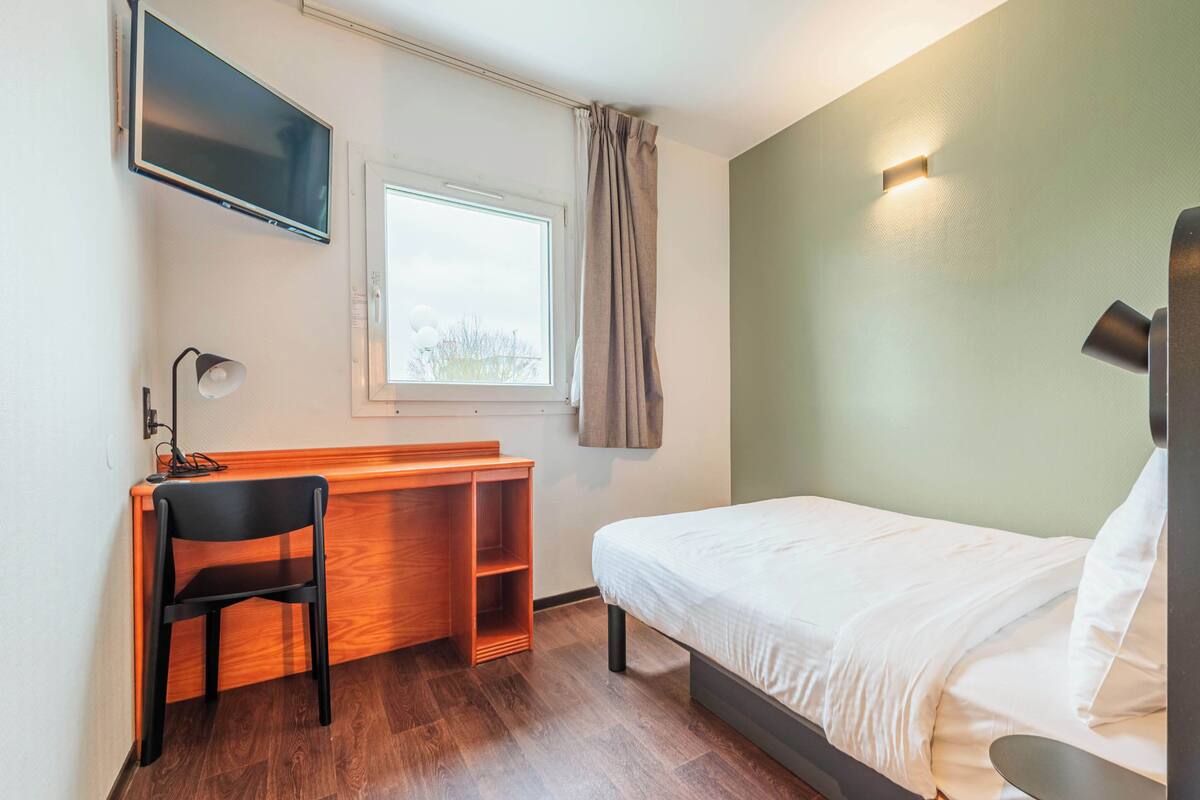 Une chambre d’hôtel comprenant un lit, un bureau avec une chaise, un téléviseur fixé au mur, une fenêtre avec des rideaux et un luminaire fixé au mur.