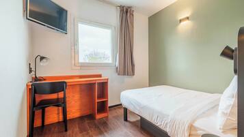 Habitación de hotel con cama, escritorio con silla, televisor montado en la pared, ventana con cortinas y lámpara de pared.