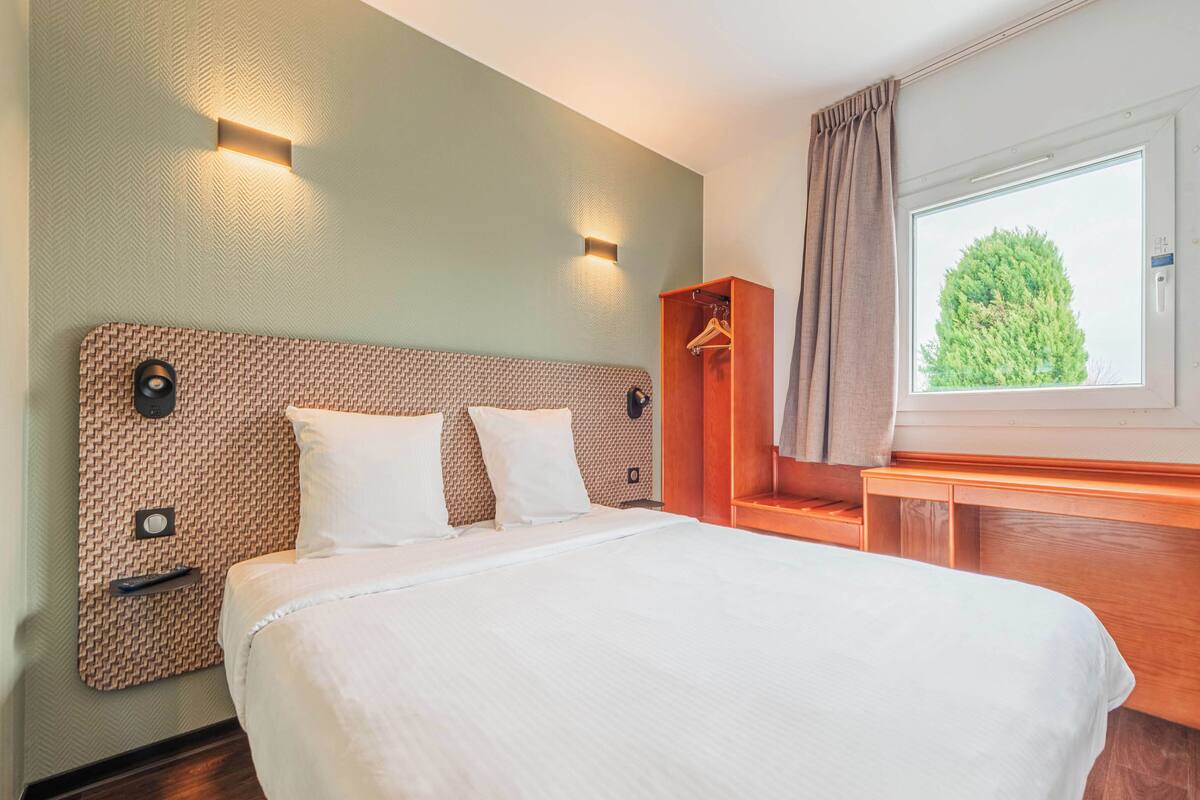 Un lit bien fait, avec une tête de lit, une table de chevet, une armoire en bois et une fenêtre avec des rideaux.