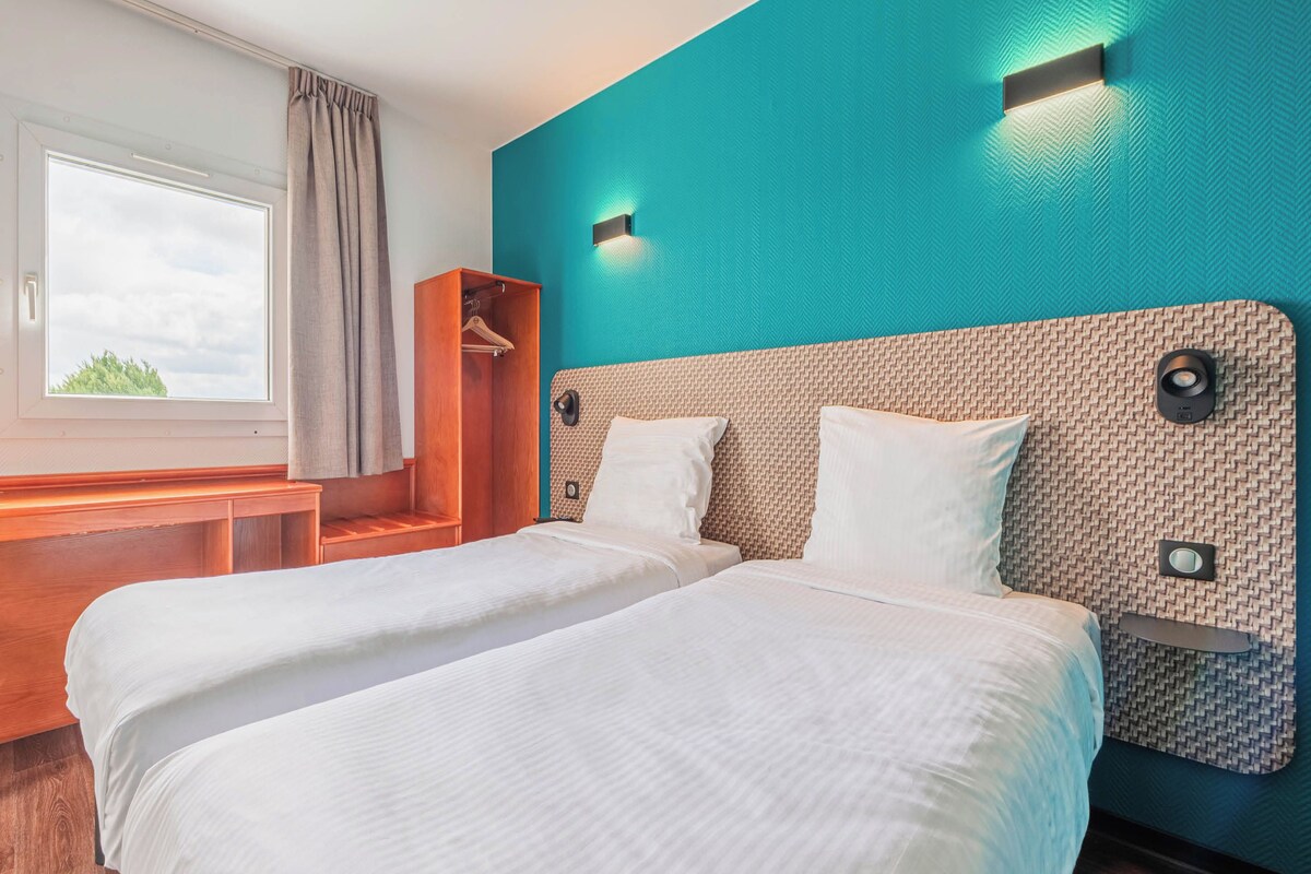 A hotel room with two beds, a wooden bench, a wardrobe, and a window with curtains.