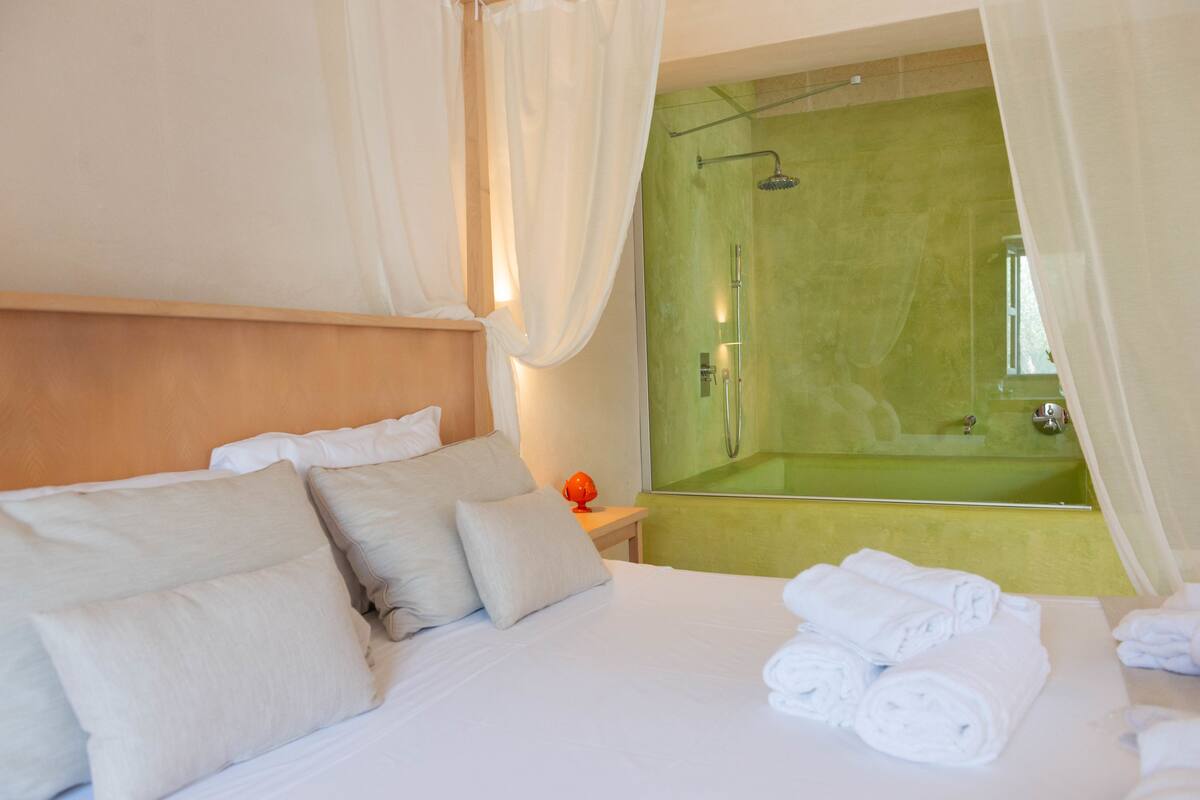 A hotel room with a bed, pillows, a nightstand, a mirror, and a green bathtub.