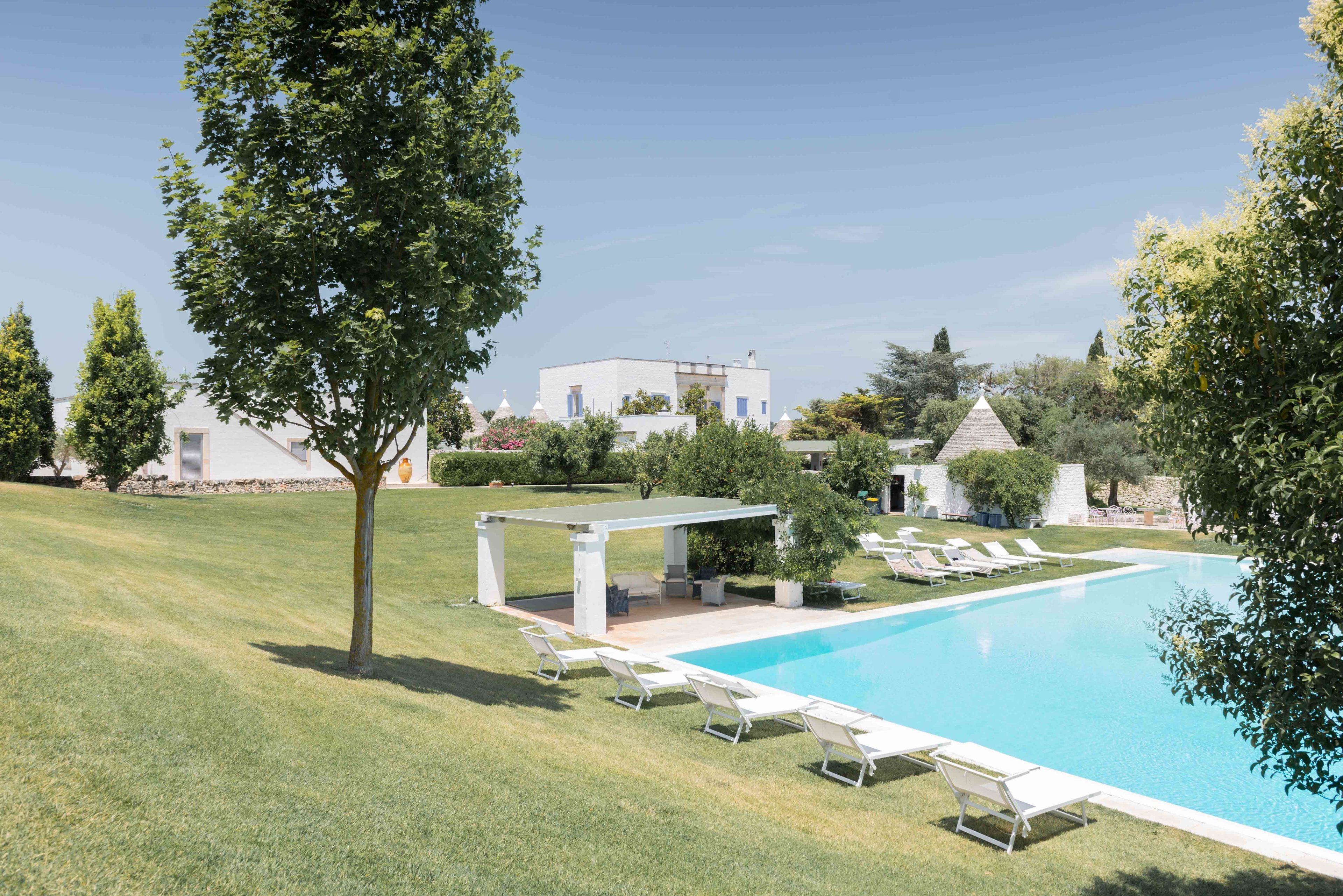 A modern pool area with a clear blue pool, surrounded by white lounge chairs and a covered seating area.