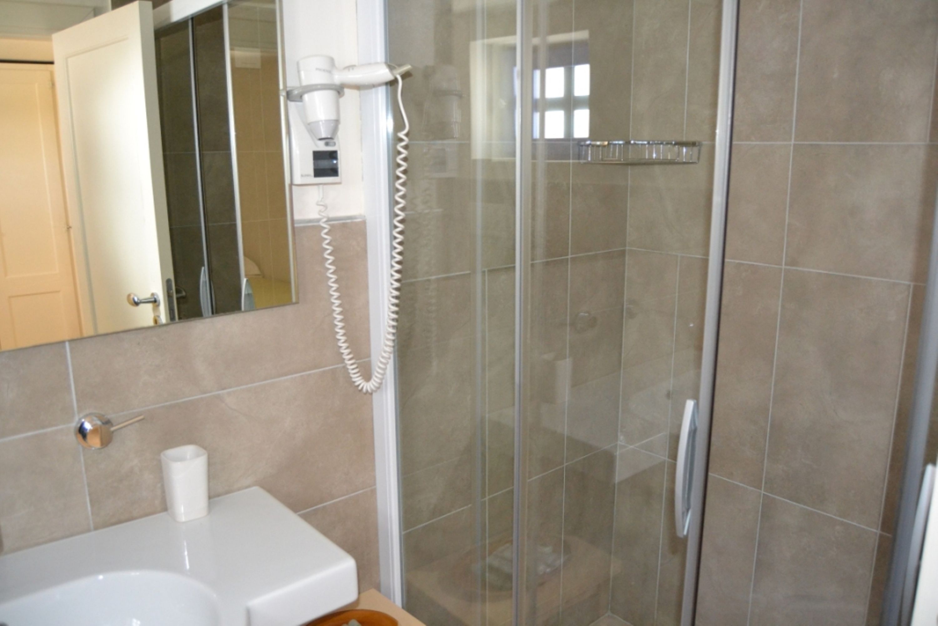 Casa de banho com cabine de duche em vidro, lavatório branco, espelho e secador de cabelo num suporte fixado à parede.