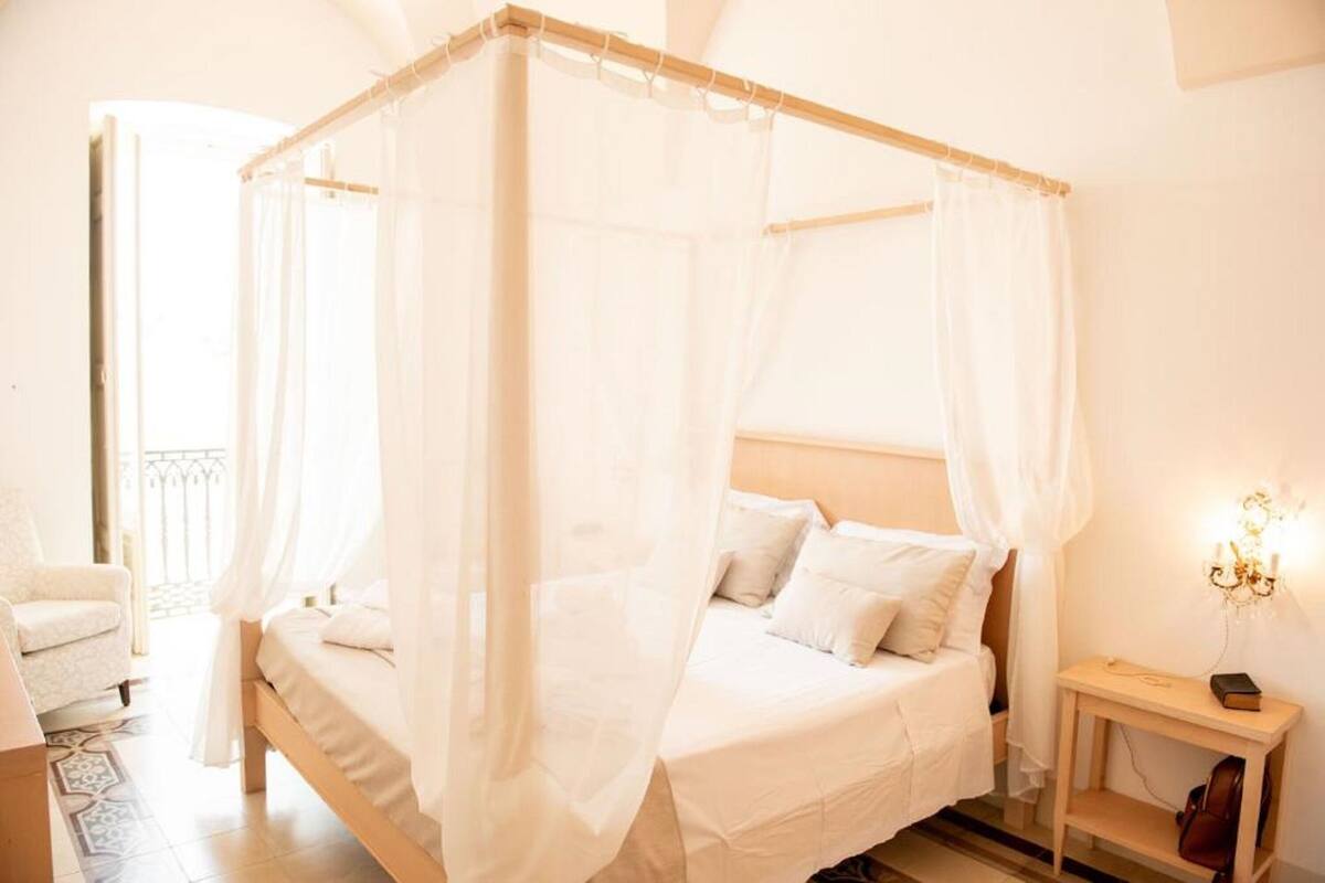 A four-poster canopy bed with white linens, a bedside table with a lamp, and a view of the balcony.