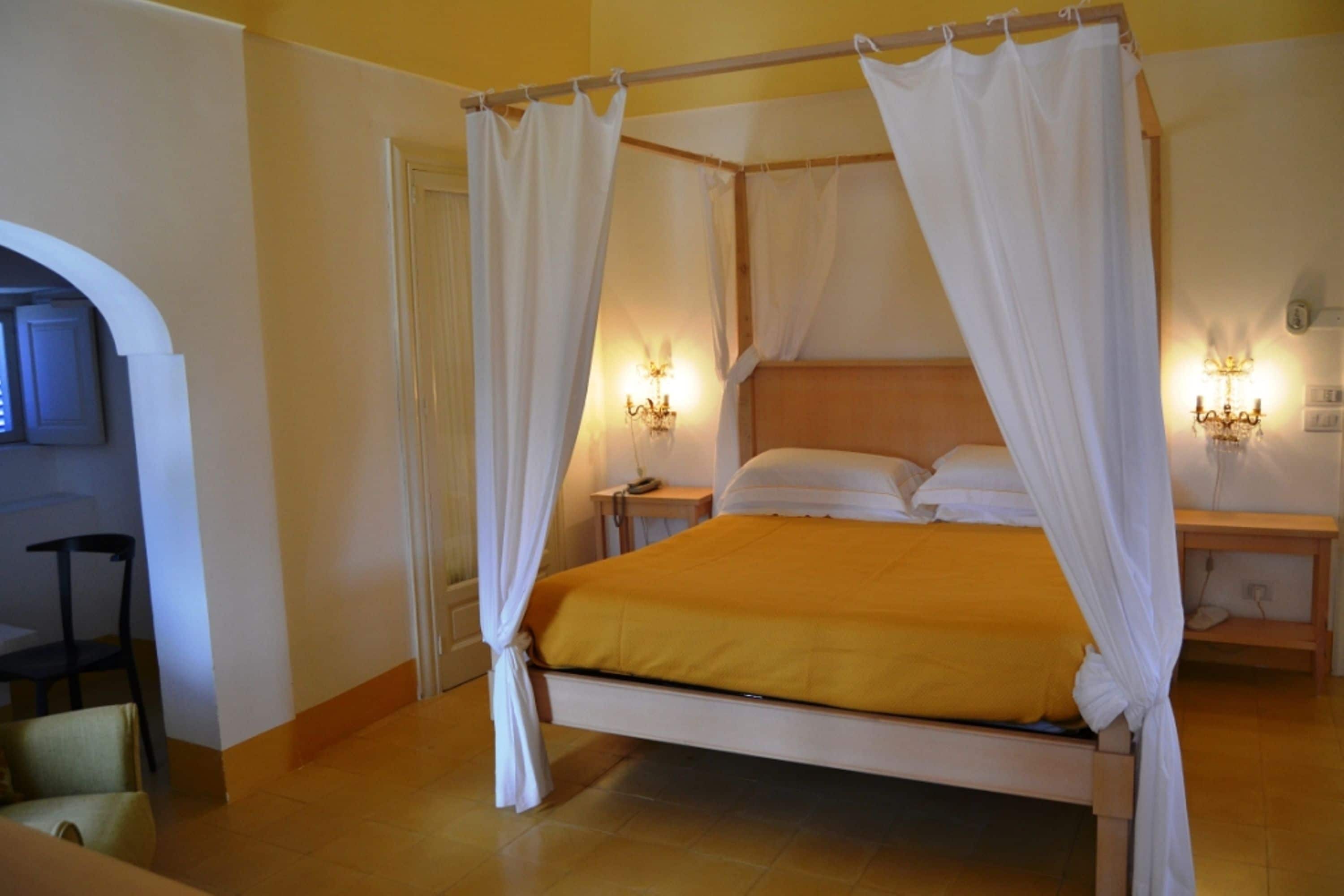 Una cama con dosel, con cortinas blancas, cabecera de madera y colcha amarilla. Hay dos mesitas de noche con lámparas.