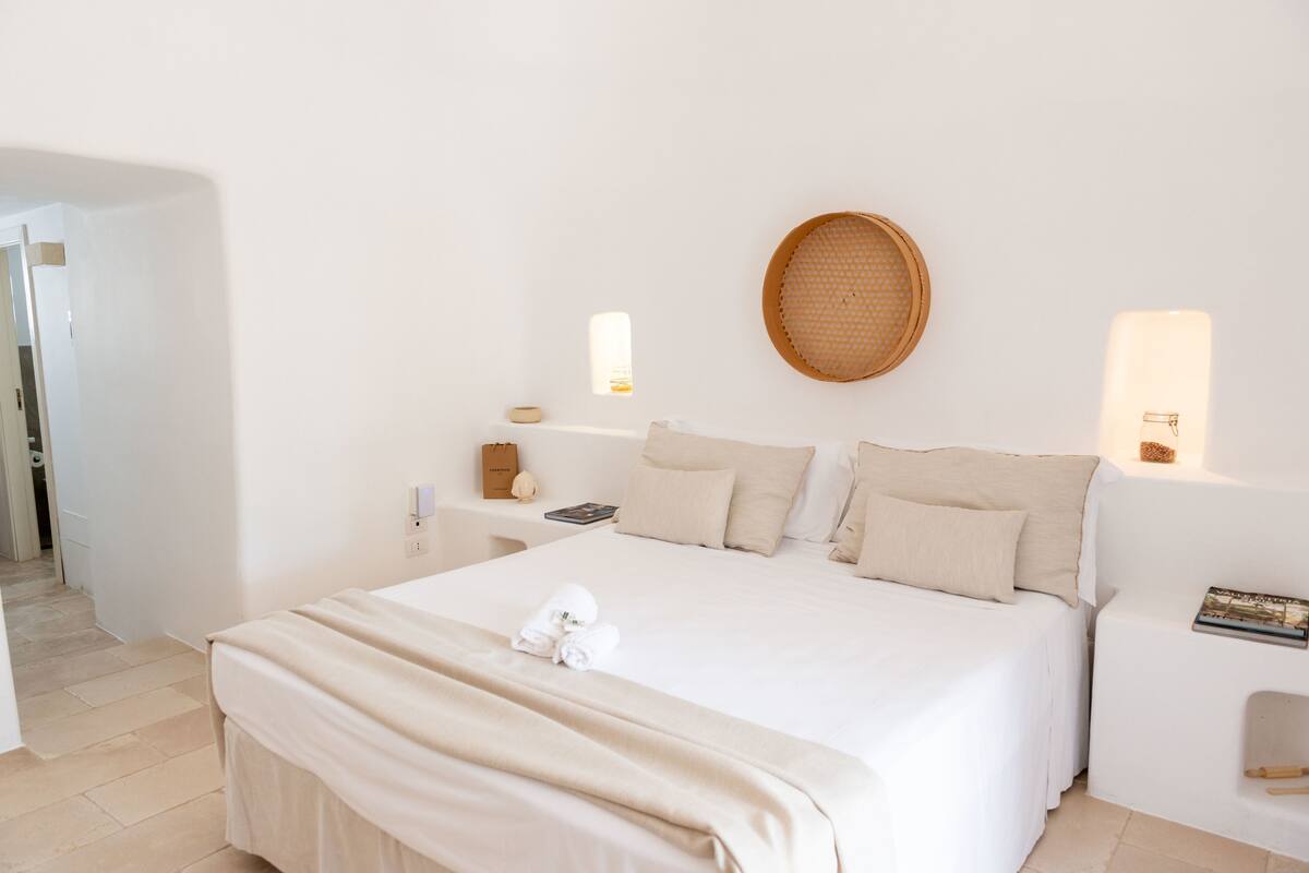 A neatly made bed with a white duvet, a round woven basket on the wall, and a small bedside table with a book and a candle.