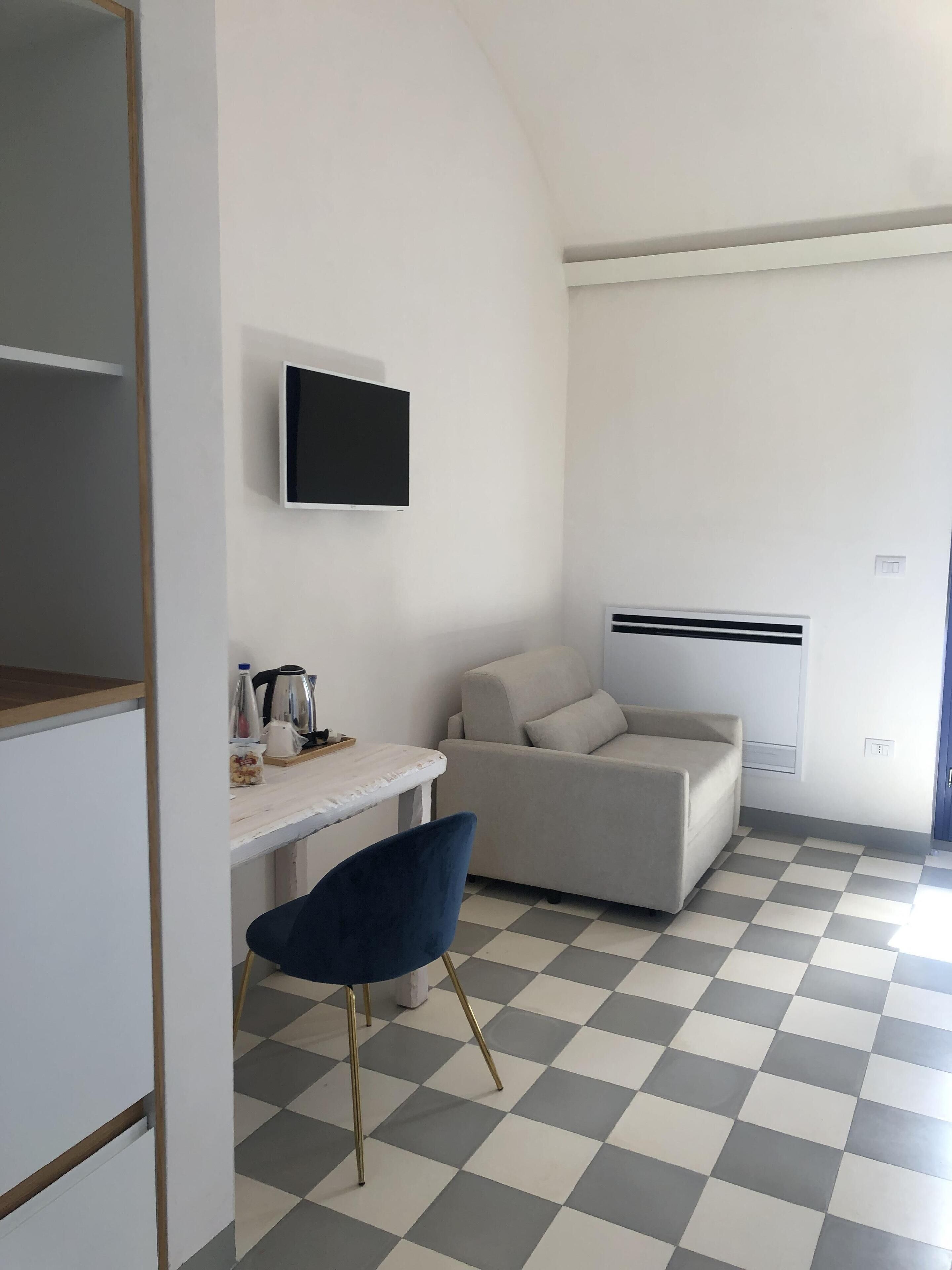 A room with a checkered floor, a white desk with a chair, a grey sofa, and a mounted TV.