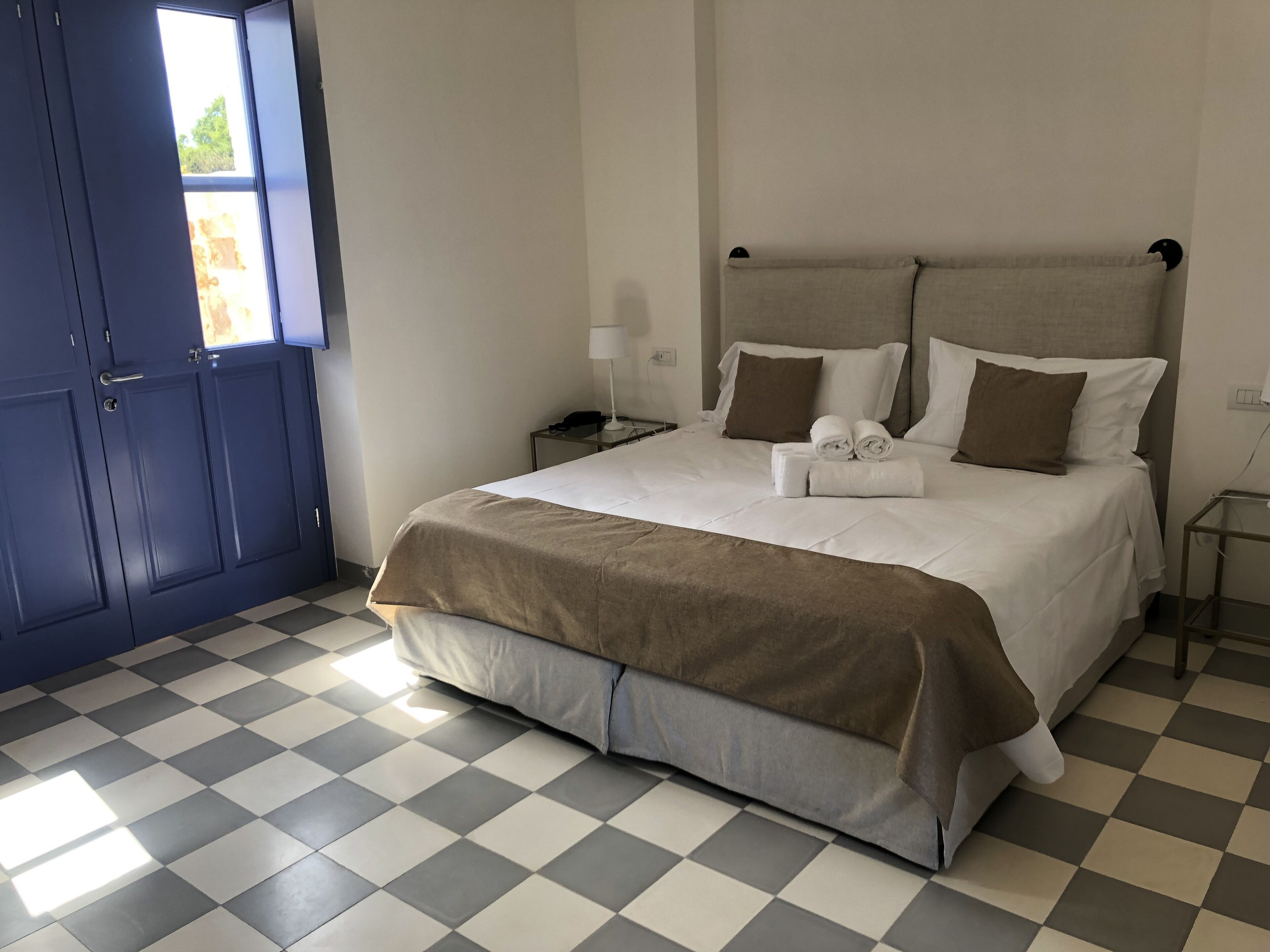 A hotel room with a checkered floor, a bed with white and brown bedding, a nightstand with a lamp, and a window with a view of greenery.