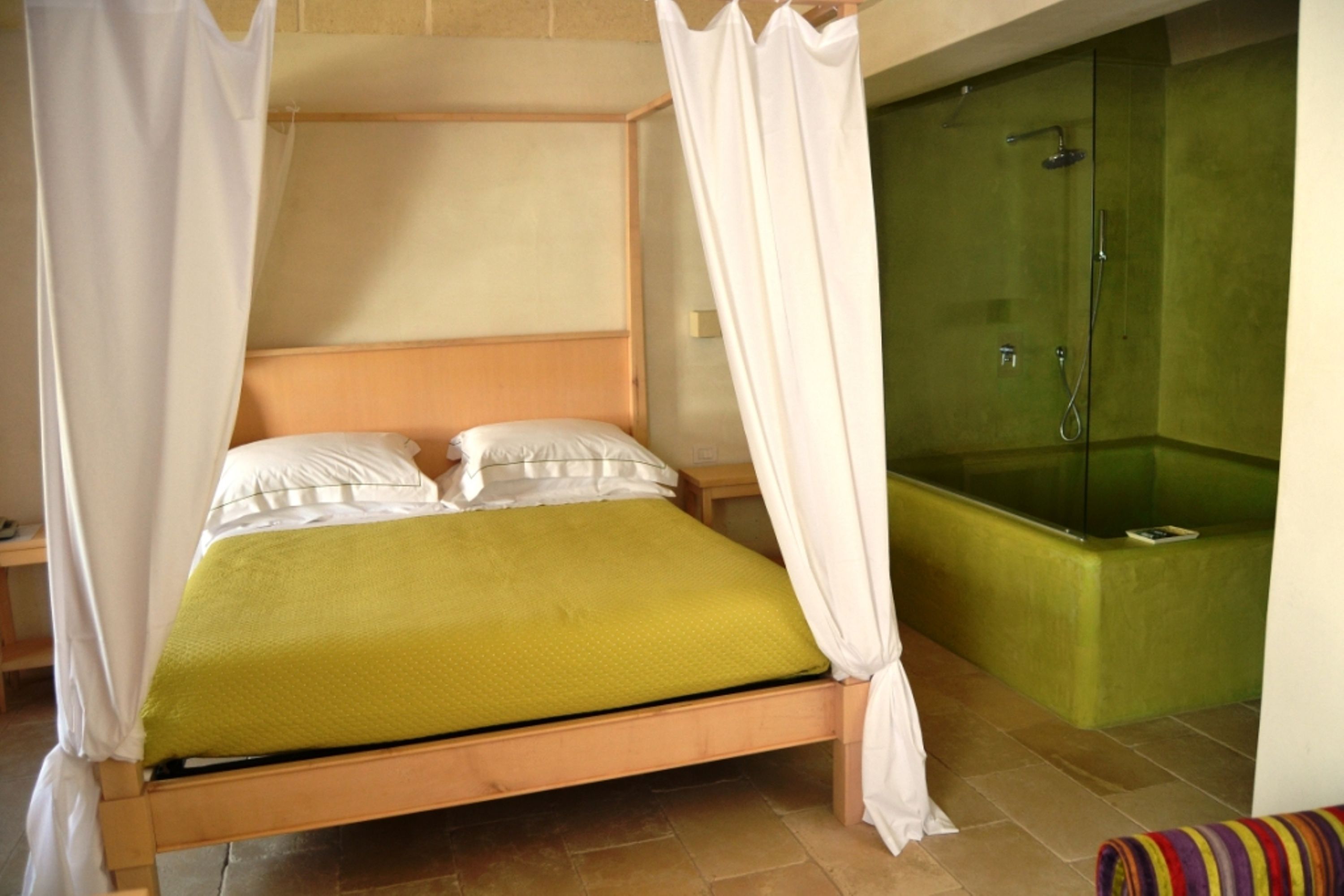 A single bed with a wooden frame and a green bedspread, a white canopy, and a green bathtub with a glass enclosure.