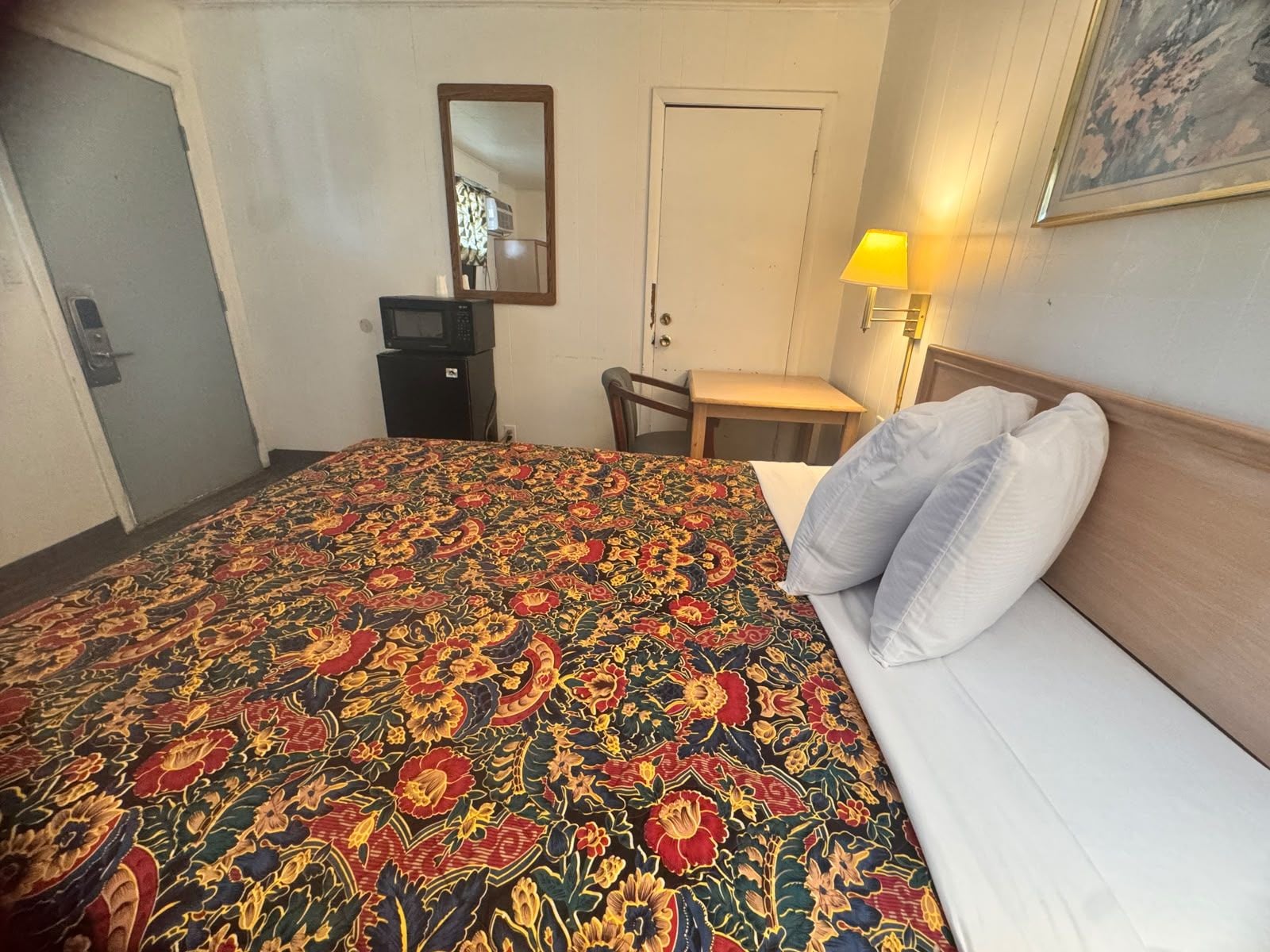 Ein Hotelzimmer mit einem Bett, einem Schreibtisch, einem Stuhl, einer Lampe, einem Spiegel und einer Tür.