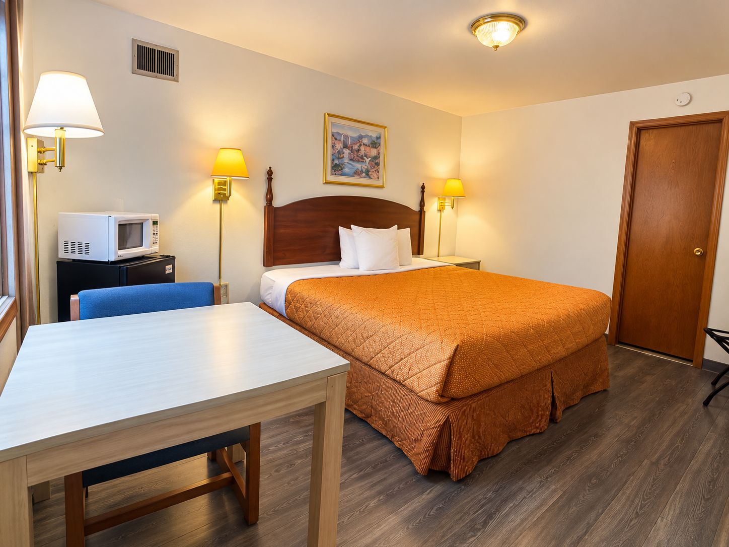 A hotel room with a bed, bedside tables, a desk, a chair, a microwave, and lamps.
